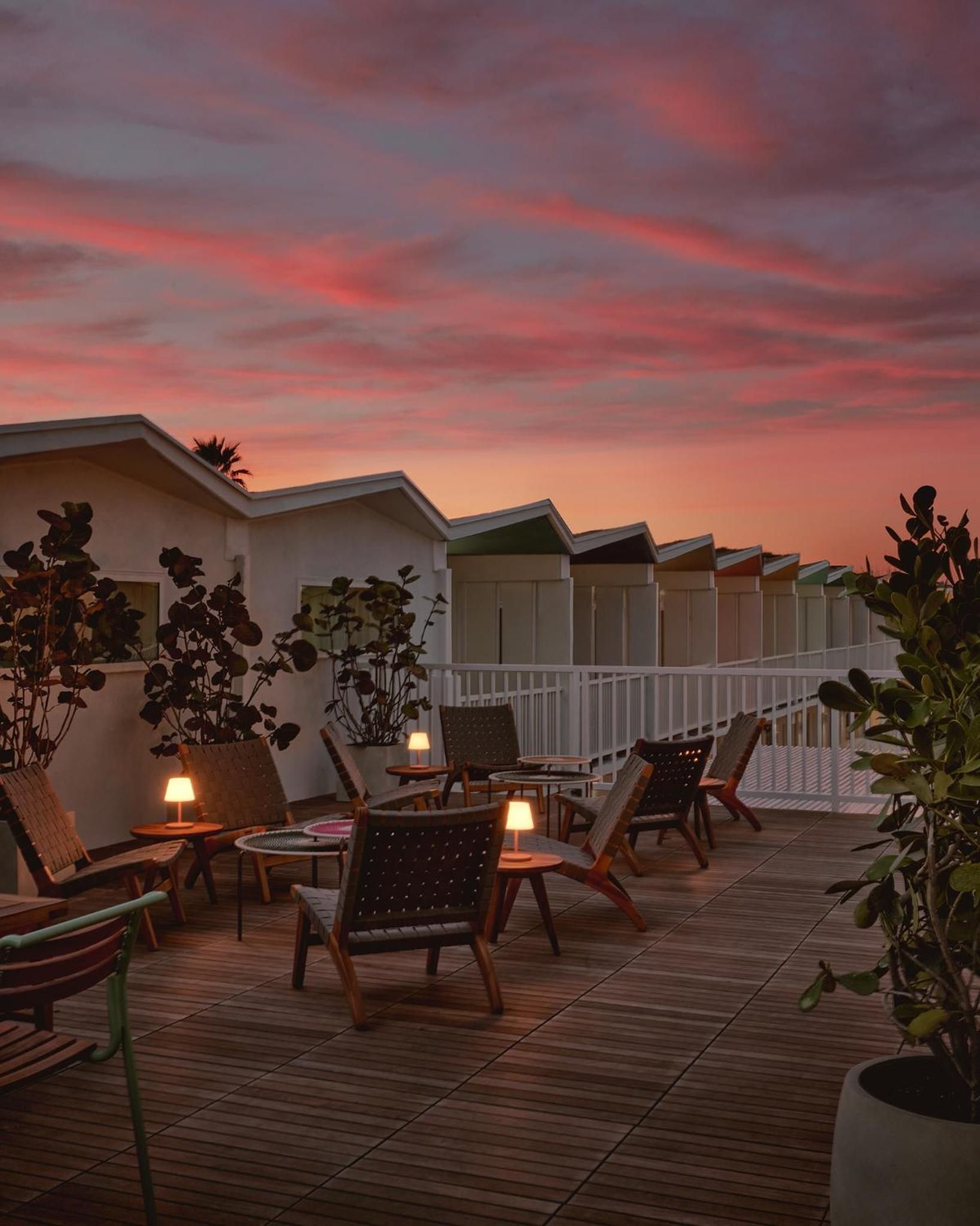 Mid-century lounge chairs and glowing table lamps create an intimate rooftop retreat as coral-streaked sunset clouds paint the sky above modern beach huts.