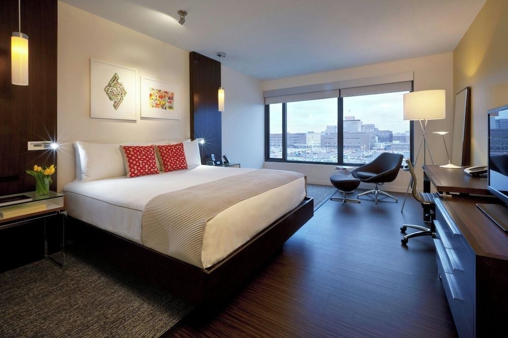 Floor-to-ceiling windows frame the urban skyline from this sleek guest room, where warm wood tones and crisp white linens create sophisticated sanctuary above the city.