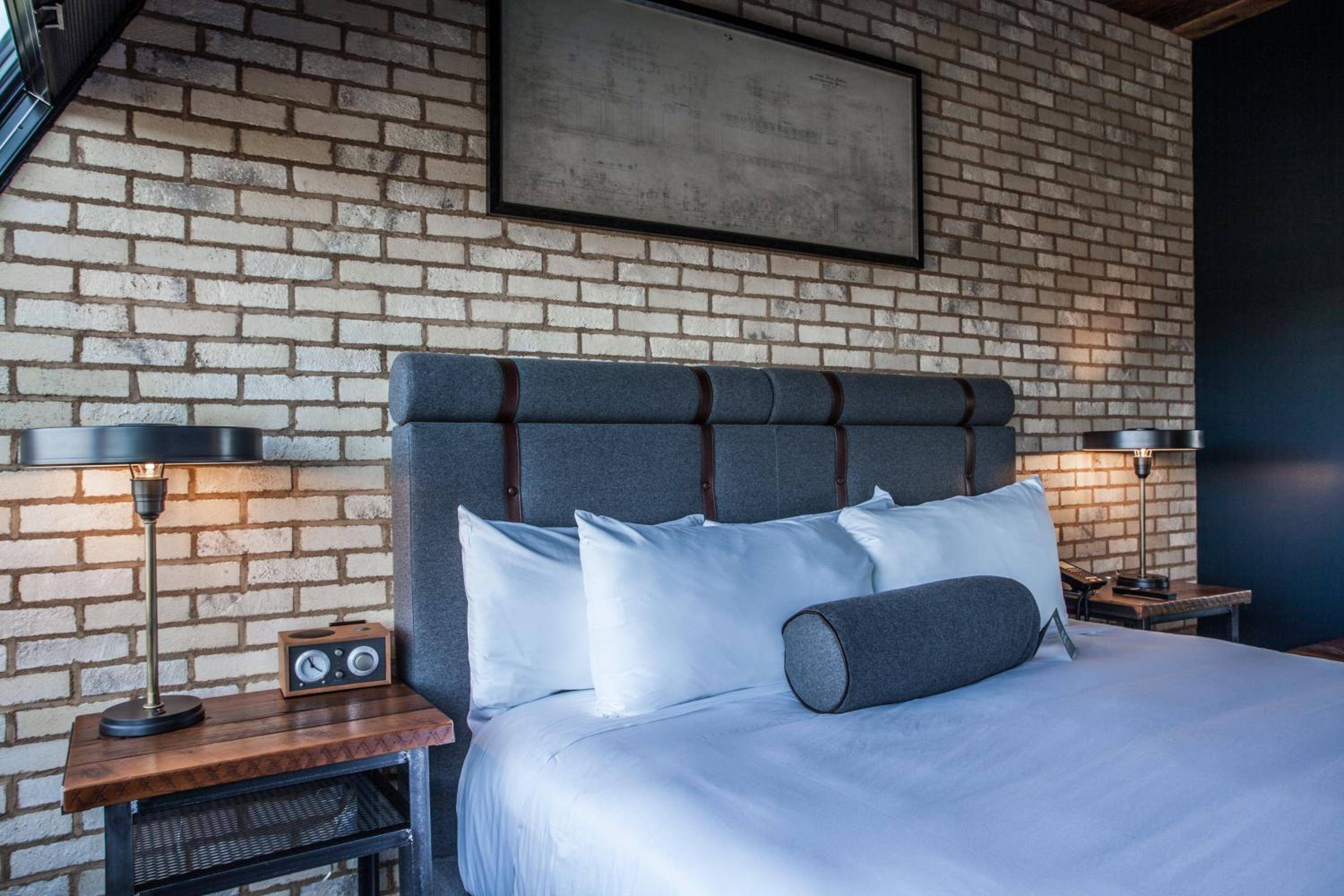 Exposed brick walls and tufted headboards create industrial-chic intimacy, while brass bedside lamps cast a warm glow over crisp white linens and vintage radio details.