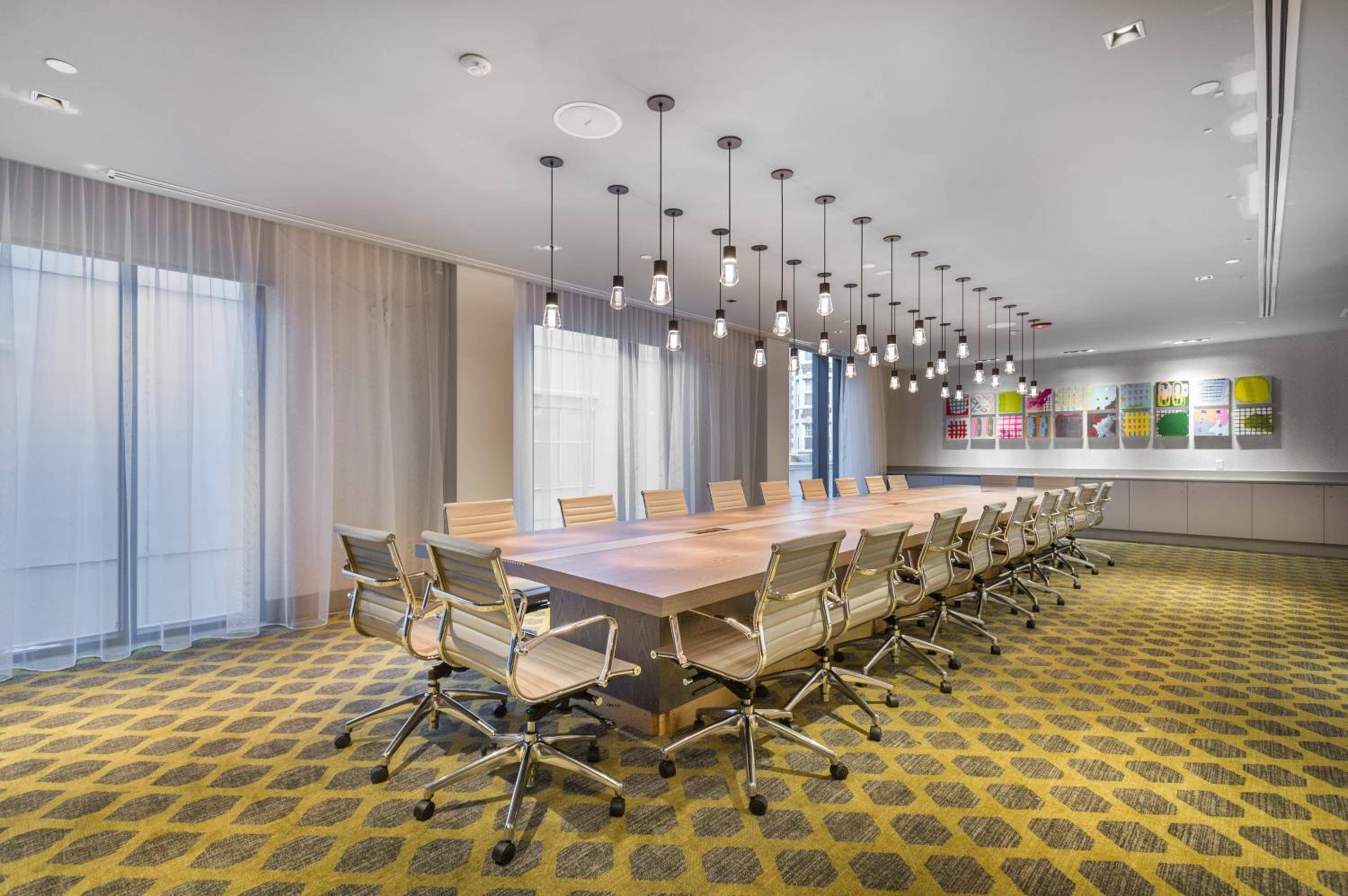 Pendant lights cascade over polished wood in this contemporary boardroom, where geometric carpeting and colorful textile samples create an unexpectedly vibrant corporate canvas.
