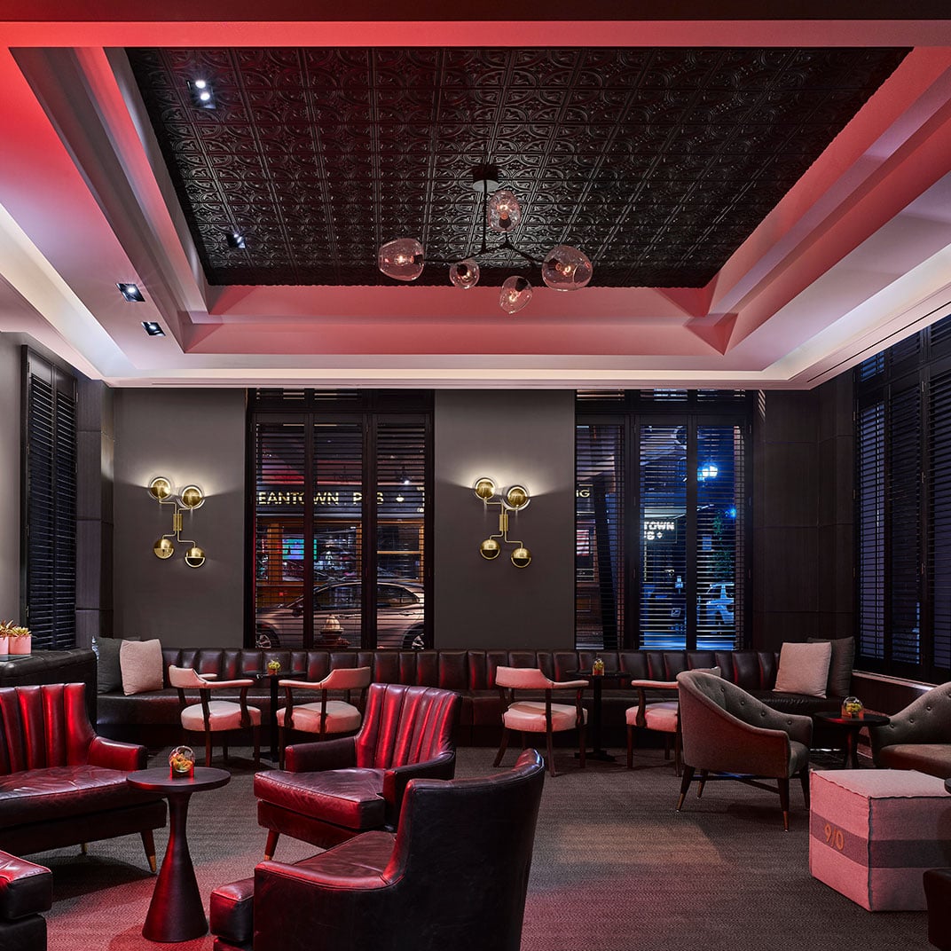 Moody crimson lighting and plush leather seating create an intimate speakeasy atmosphere, where ornate tin ceilings frame golden bubble chandeliers above dark wood furnishings.