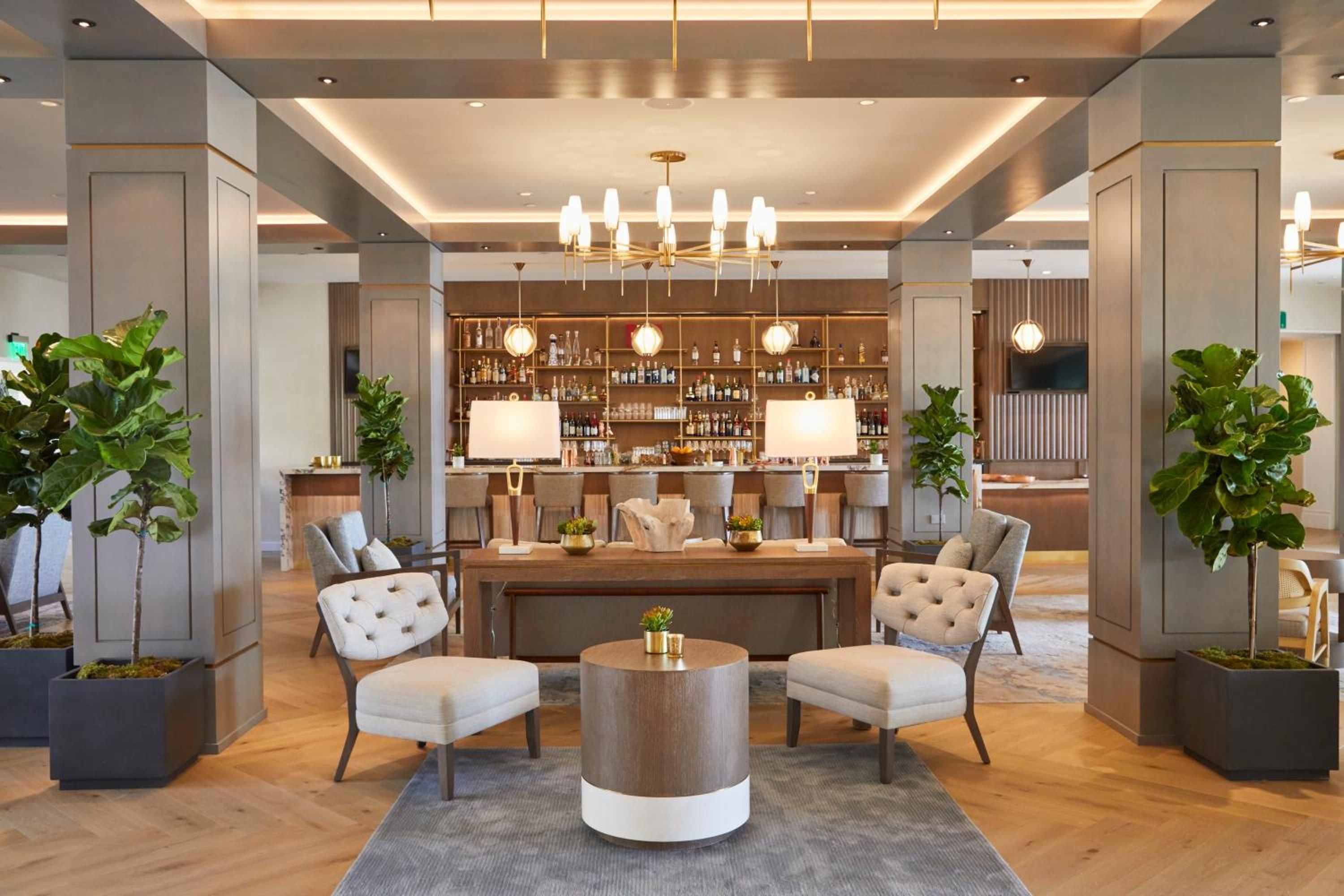 Warm wood tones and sage-green columns frame the sophisticated lobby lounge, where fiddle-leaf figs cast gentle shadows and golden pendant lights create intimate conversation nooks.