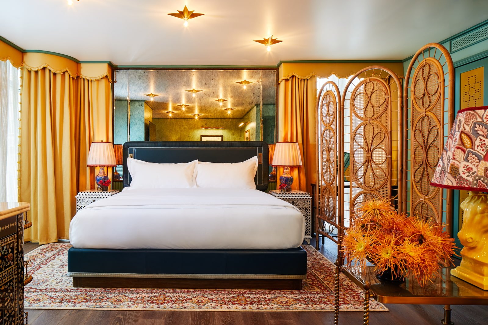 Golden curtains frame crisp white linens while star-shaped ceiling lights cast warm shadows across ornate screens and vibrant orange florals in this richly layered retreat.