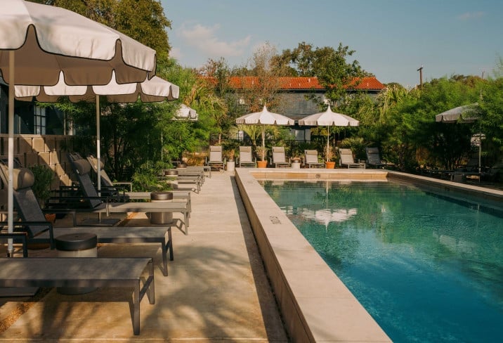 Sunlight filters through leafy canopies onto polished concrete, where sleek loungers and vintage umbrellas frame the sapphire pool in understated luxury.