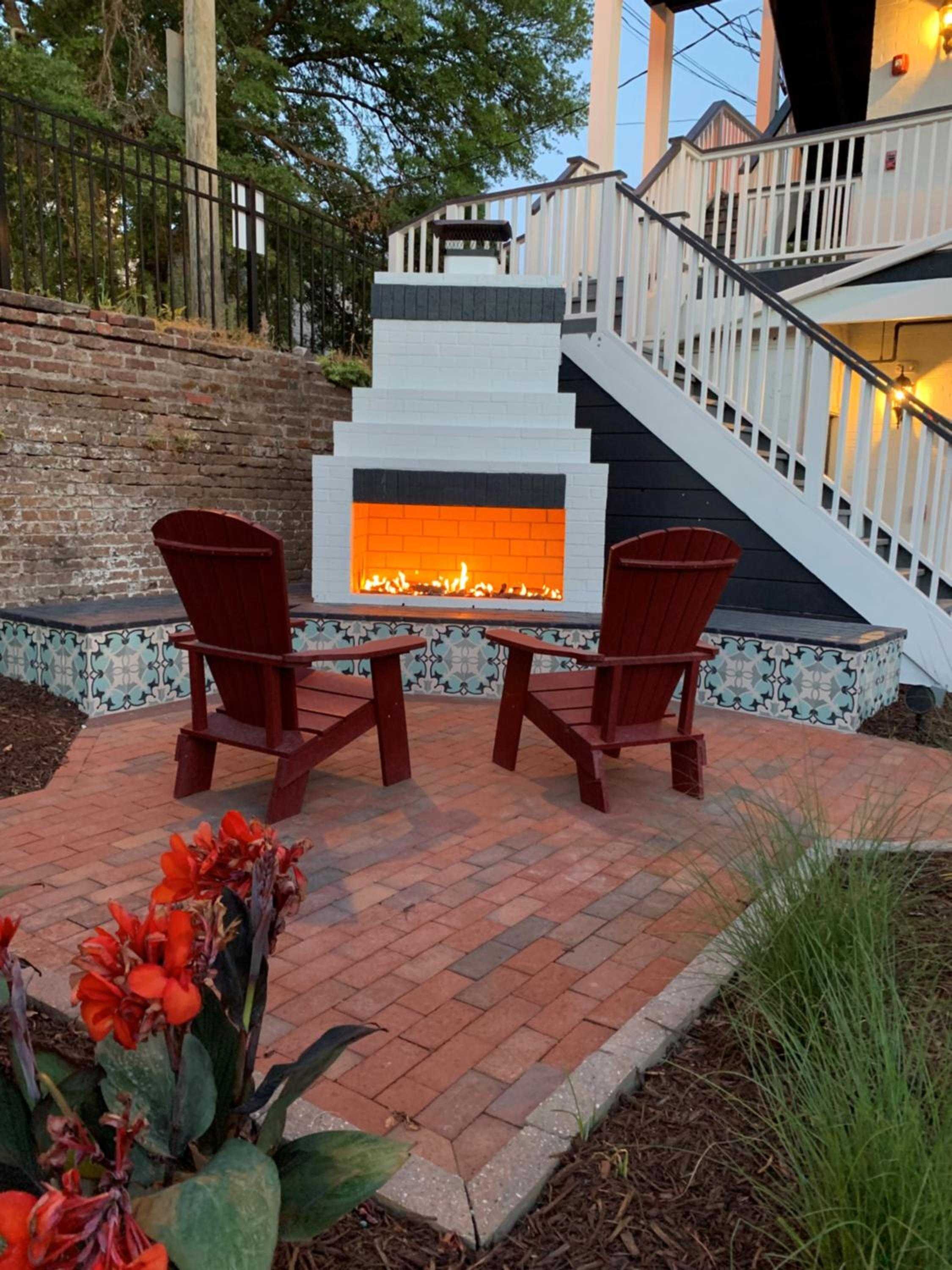 Red Adirondack chairs frame a contemporary outdoor fireplace beneath white deck stairs, where patterned tiles and warm brick create an inviting evening retreat.