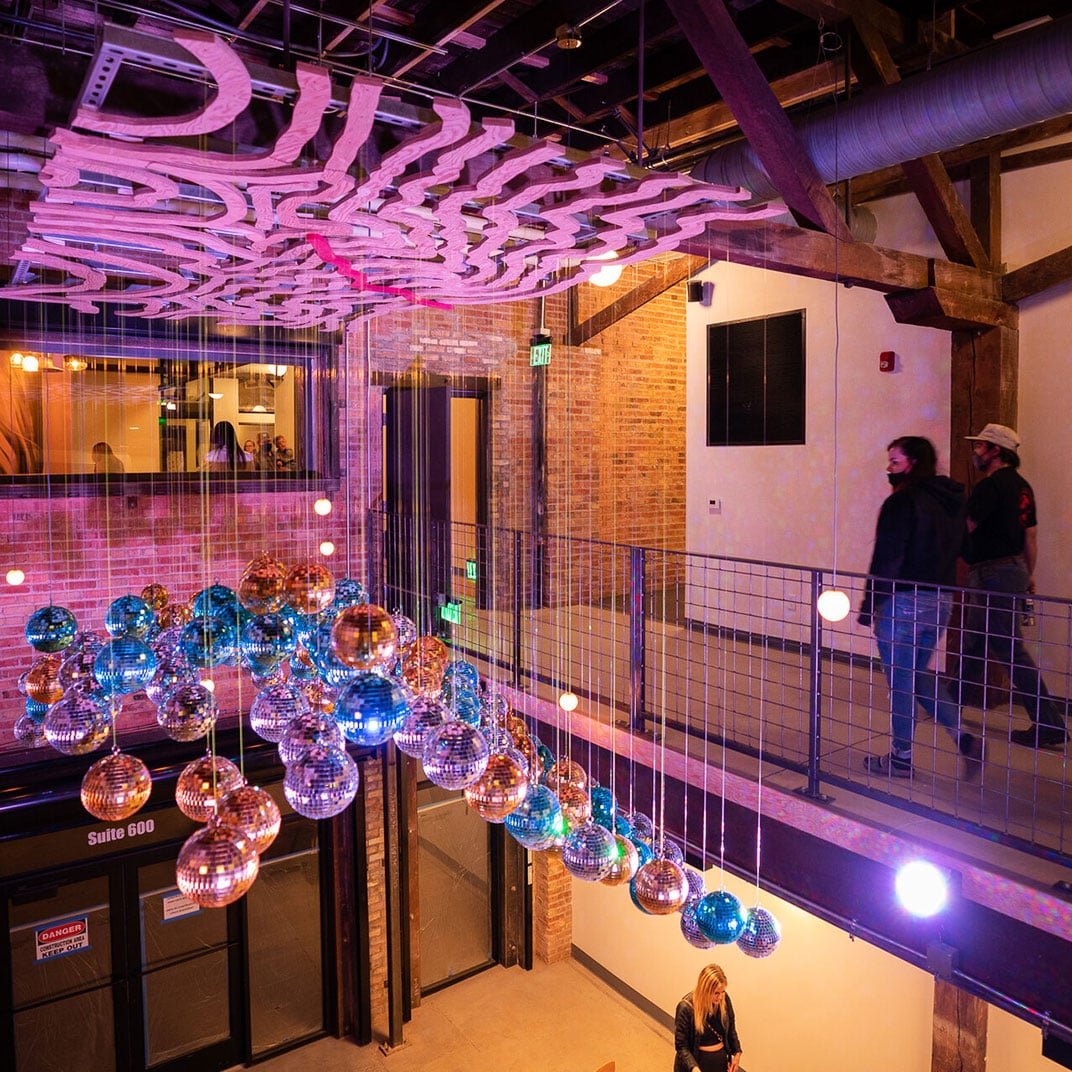 Suspended disco balls cascade through the industrial-chic atrium, their mirrored surfaces catching purple LED light while guests marvel from the exposed-beam mezzanine above.