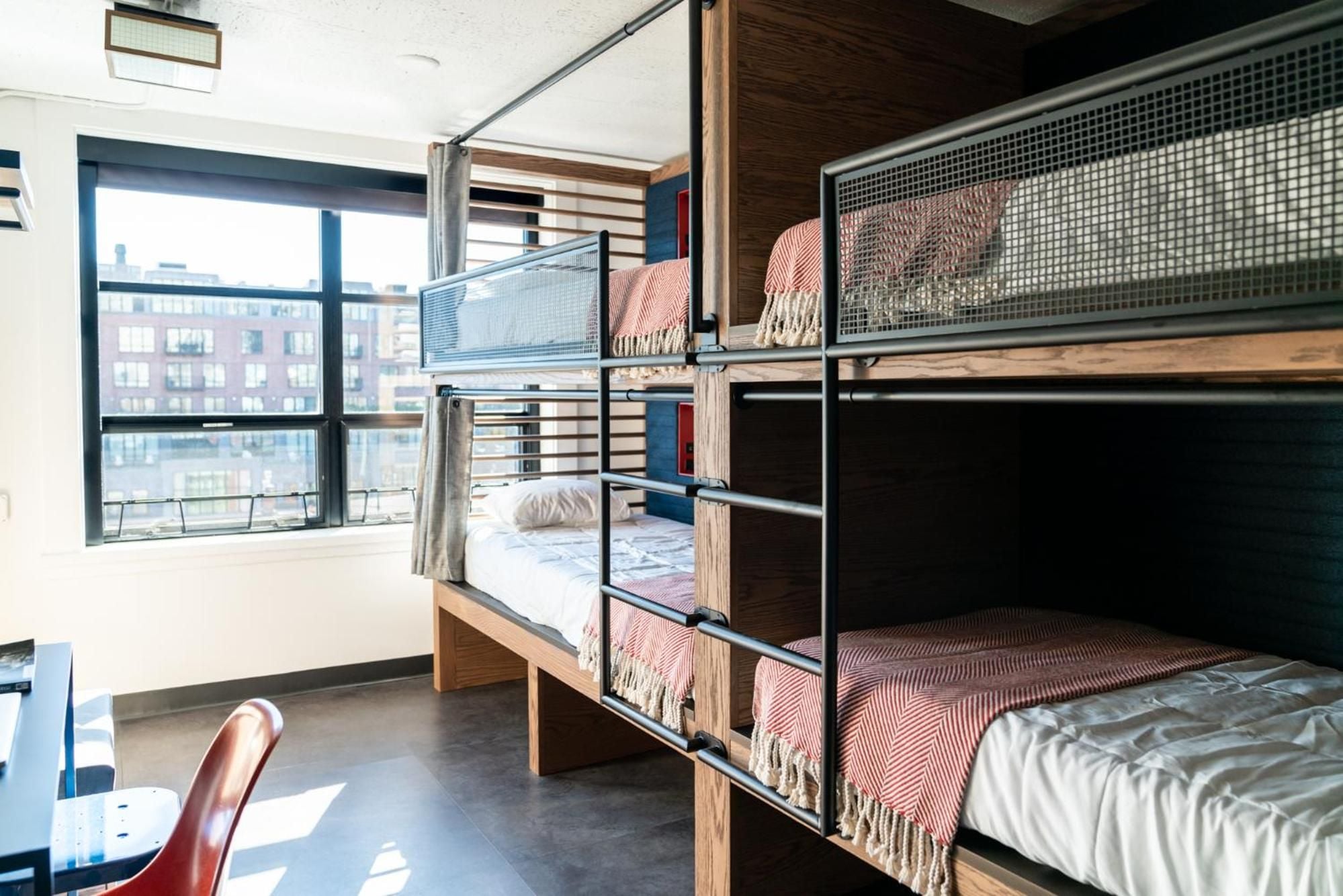 Modern bunk beds with geometric mesh panels and coral-striped throws create an unexpectedly stylish shared accommodation, while floor-to-ceiling windows frame Boston's urban landscape beyond.