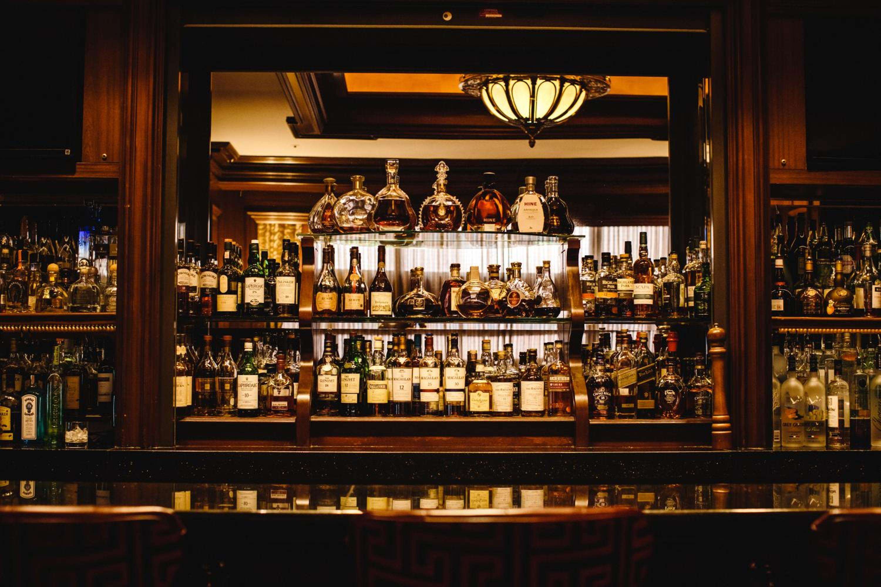 Amber light bathes an impressive whiskey collection behind polished wood and glass, where premium spirits create a sophisticated backdrop for evening libations.