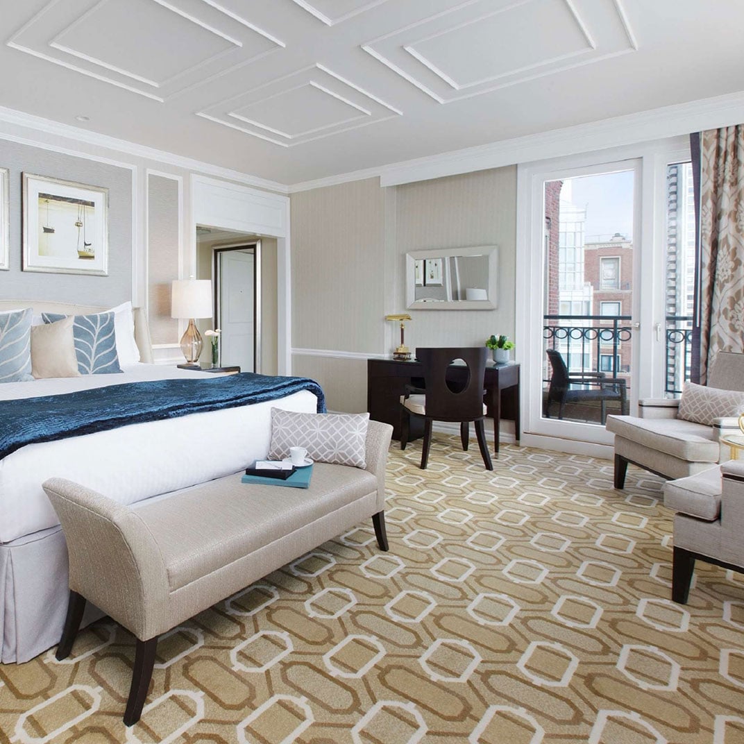 Geometric carpets and coffered ceilings create sophisticated layers in this harbor-view suite, where French doors frame Boston's historic brick architecture beyond the wrought-iron balcony.