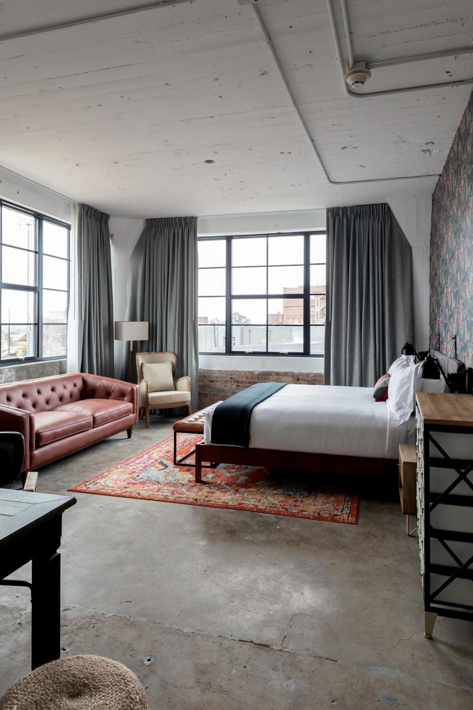 Industrial elegance meets Southern comfort in this loft-style suite, where exposed concrete ceilings frame vintage Persian rugs and rich leather furnishings beneath soaring warehouse windows.
