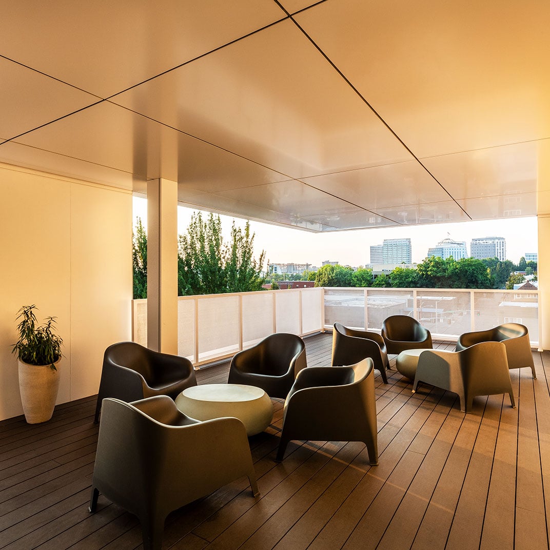 Sculptural armchairs cluster beneath a dramatic cantilevered ceiling on Jupiter NEXT's rooftop terrace, where warm timber decking frames panoramic cityscape views through contemporary design.
