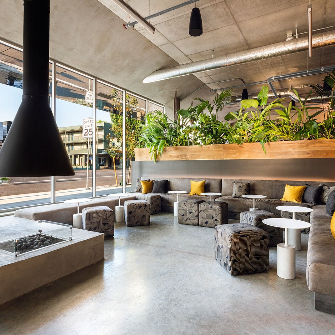 Polished concrete floors meet industrial-chic design in this inviting lounge, where geometric ottomans cluster around sleek tables beneath soaring concrete ceilings and abundant greenery.
