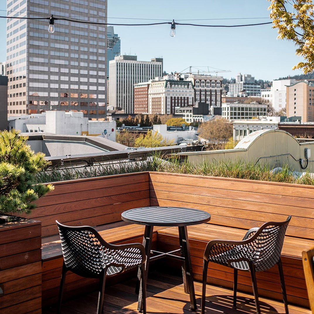 String lights dangle above weathered wood seating as Portland's skyline stretches beyond this intimate rooftop retreat, where city energy meets elevated calm.