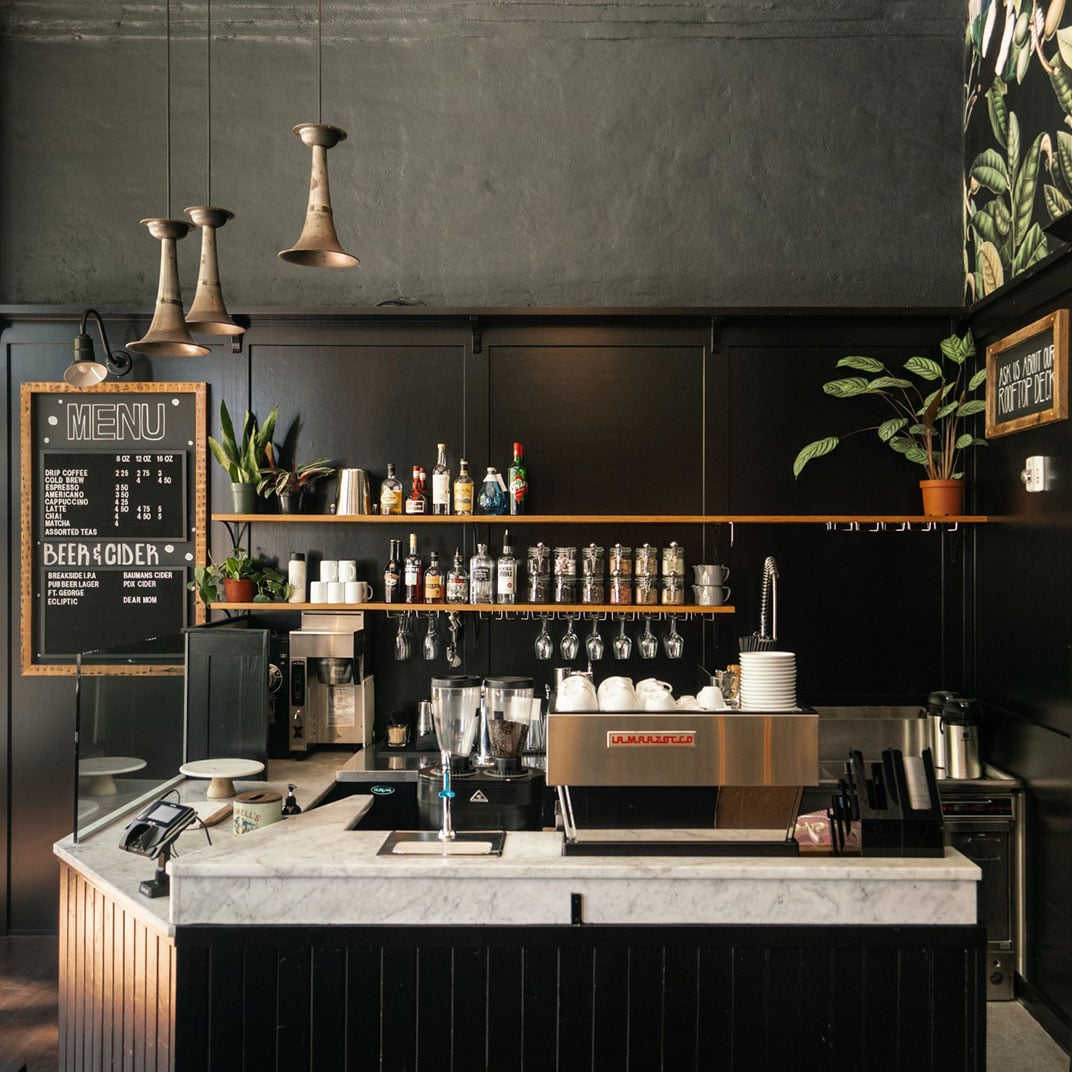 Copper pendant lights cast warm glows over the marble-topped coffee bar, where botanical wallpaper and vintage brass fixtures create an intimate, artisanal atmosphere.