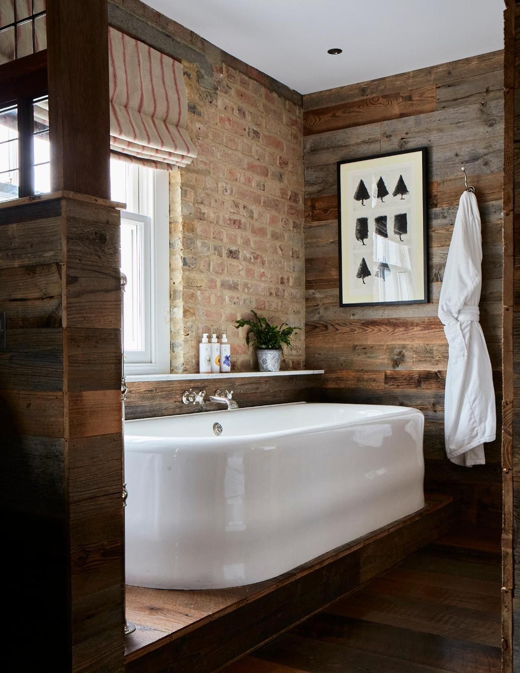 Reclaimed wood planks and exposed brick create a cozy cocoon around the freestanding tub, where botanical prints and striped Roman shades add countryside charm.