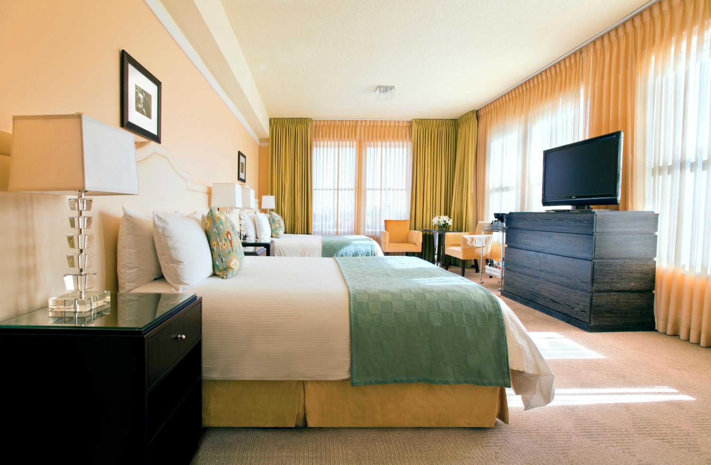 Warm amber light filters through flowing curtains, illuminating sage green accents and polished dark wood furniture in this elegantly appointed guest room.