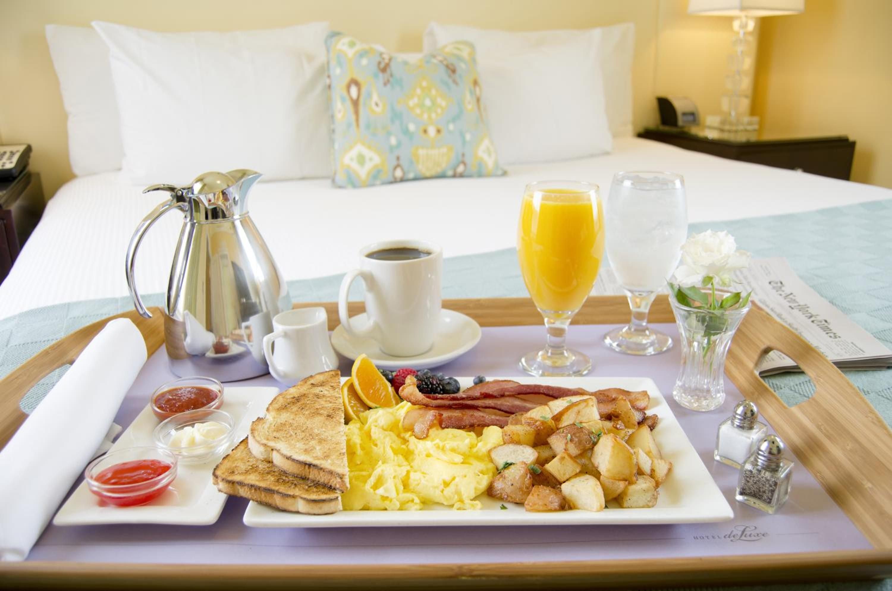 Morning sunlight streams across crisp white linens as breakfast arrives on a wooden tray, complete with fluffy scrambled eggs, crispy bacon, and fresh orange juice.