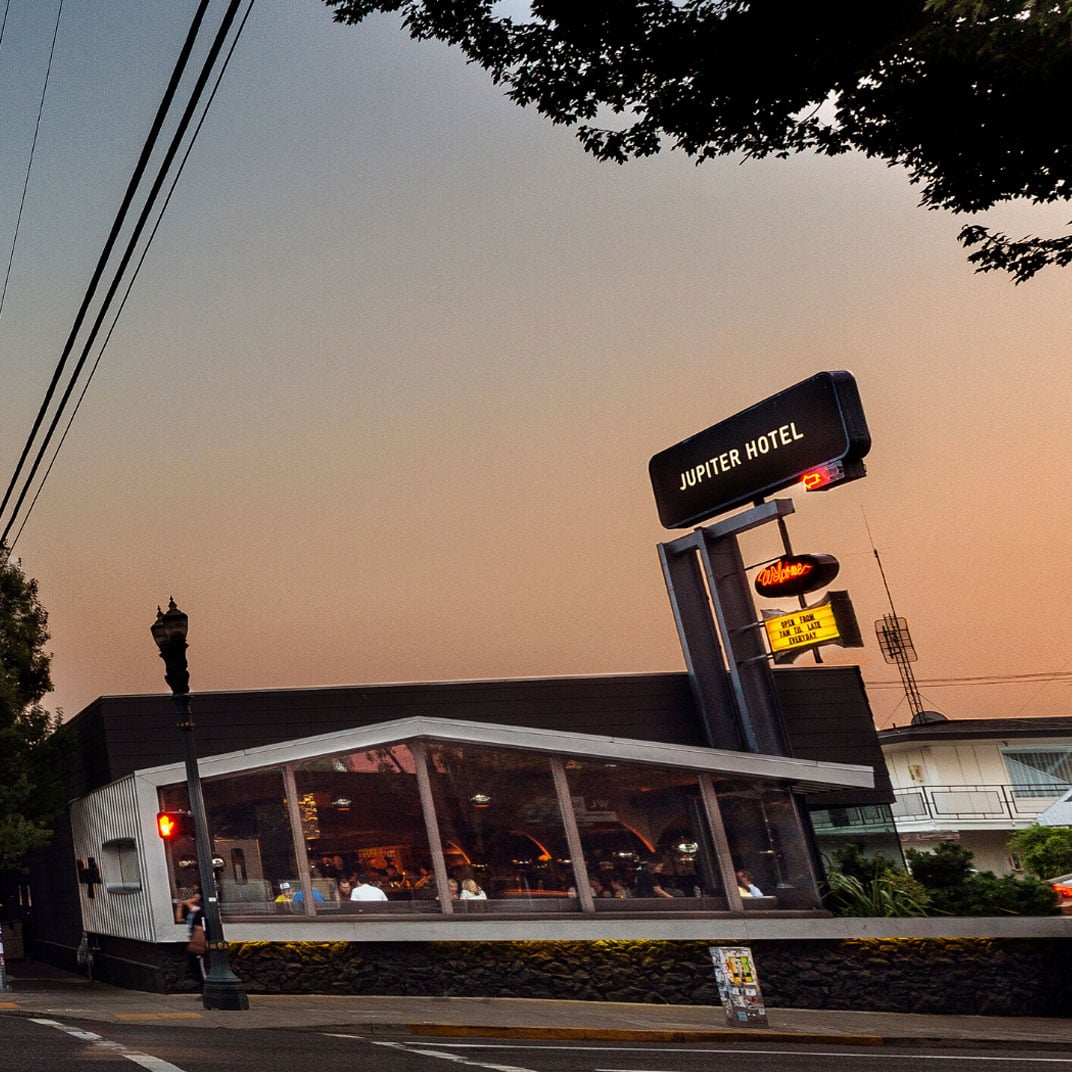 Mid-century charm radiates from Jupiter Hotel's neon-lit diner as twilight bathes the Portland sky in peachy hues, promising retro comfort and late-night nostalgia.