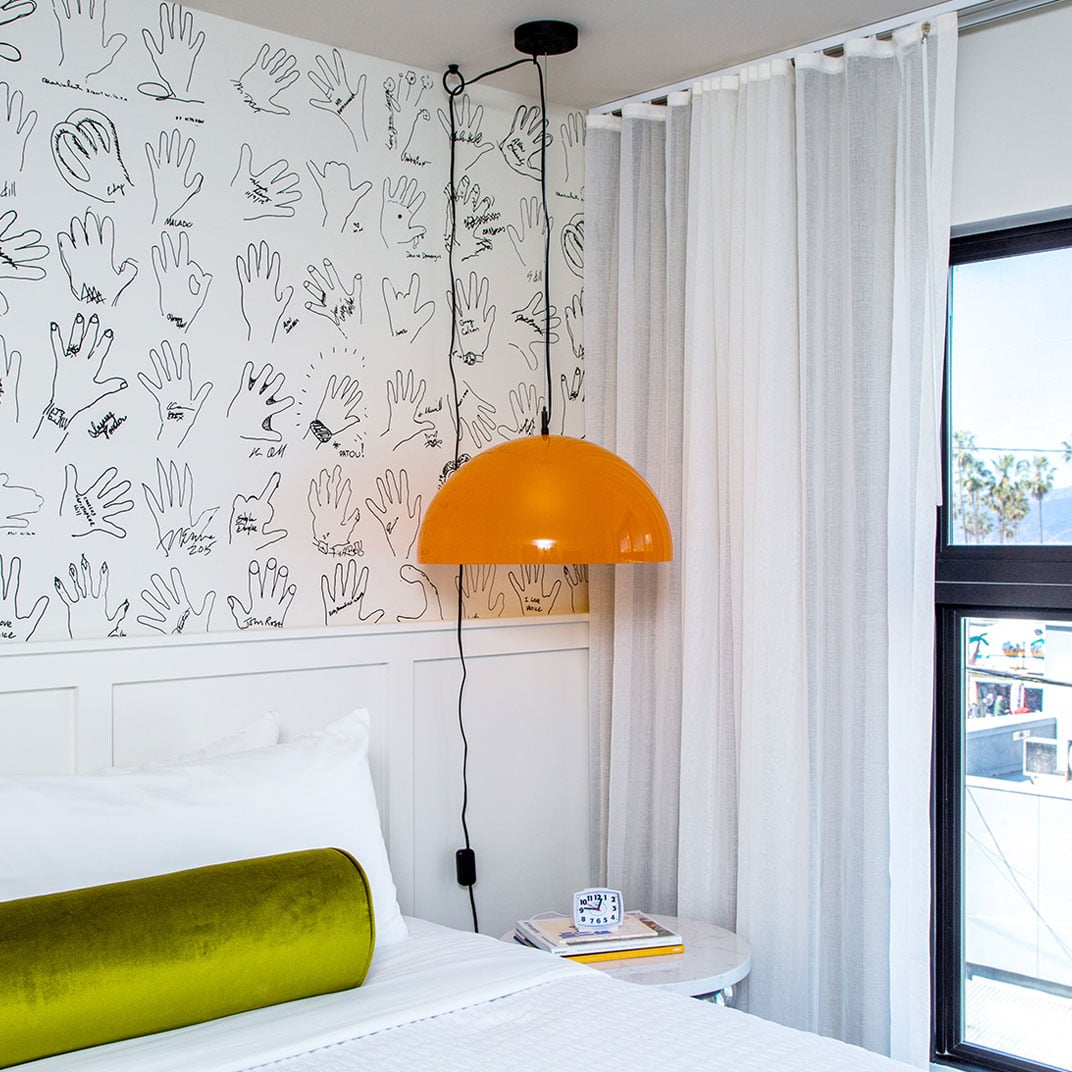 Whimsical hand-drawn wallpaper and a vibrant orange pendant lamp create playful sophistication above crisp white linens and a chartreuse bolster pillow.