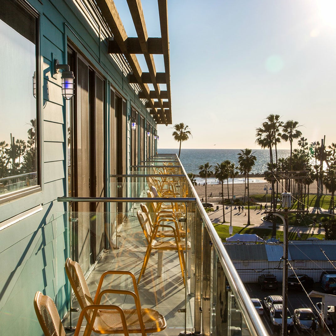 Golden hour bathes the oceanfront balcony in warm light, where modern chairs line the glass-railed terrace overlooking Venice Beach's iconic palms and endless Pacific horizon.