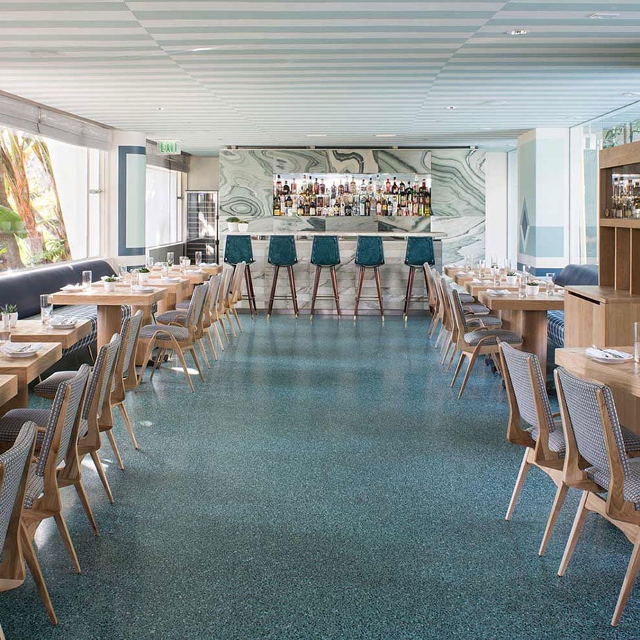 Aquamarine terrazzo floors and natural stone complement warm wood furnishings, while teal bar stools add coastal charm to this inviting dining space.
