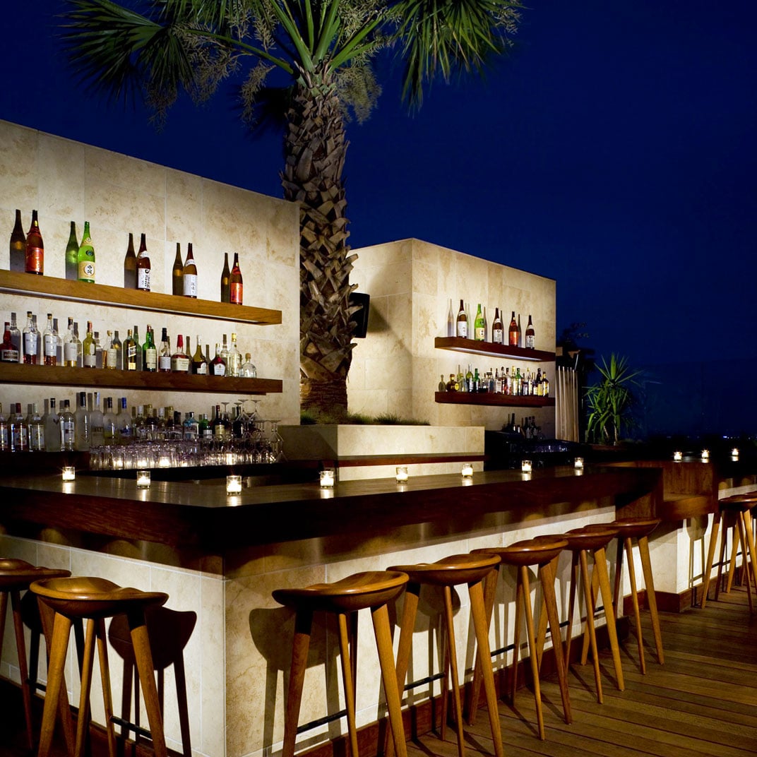 Warm candlelight dances across polished wood surfaces while palm fronds frame the evening sky at this sophisticated rooftop bar retreat.