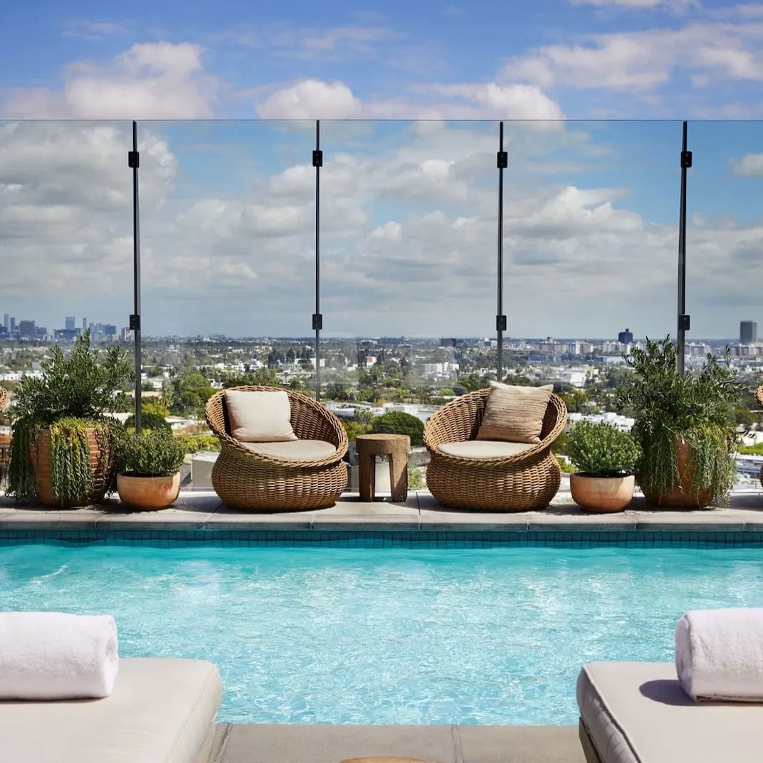 Woven rattan cocoon chairs frame the rooftop infinity pool, where crystalline waters mirror Los Angeles sprawling beneath billowing clouds and endless sky.