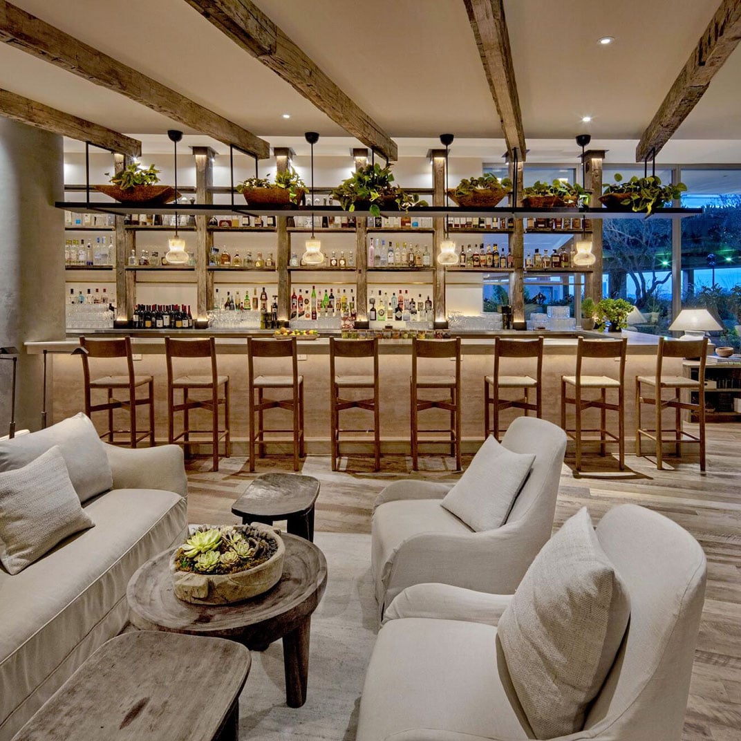 Exposed wood beams frame this sophisticated bar where leather stools line marble counters, pendant lights cast warm glows, and lush greenery softens industrial edges.