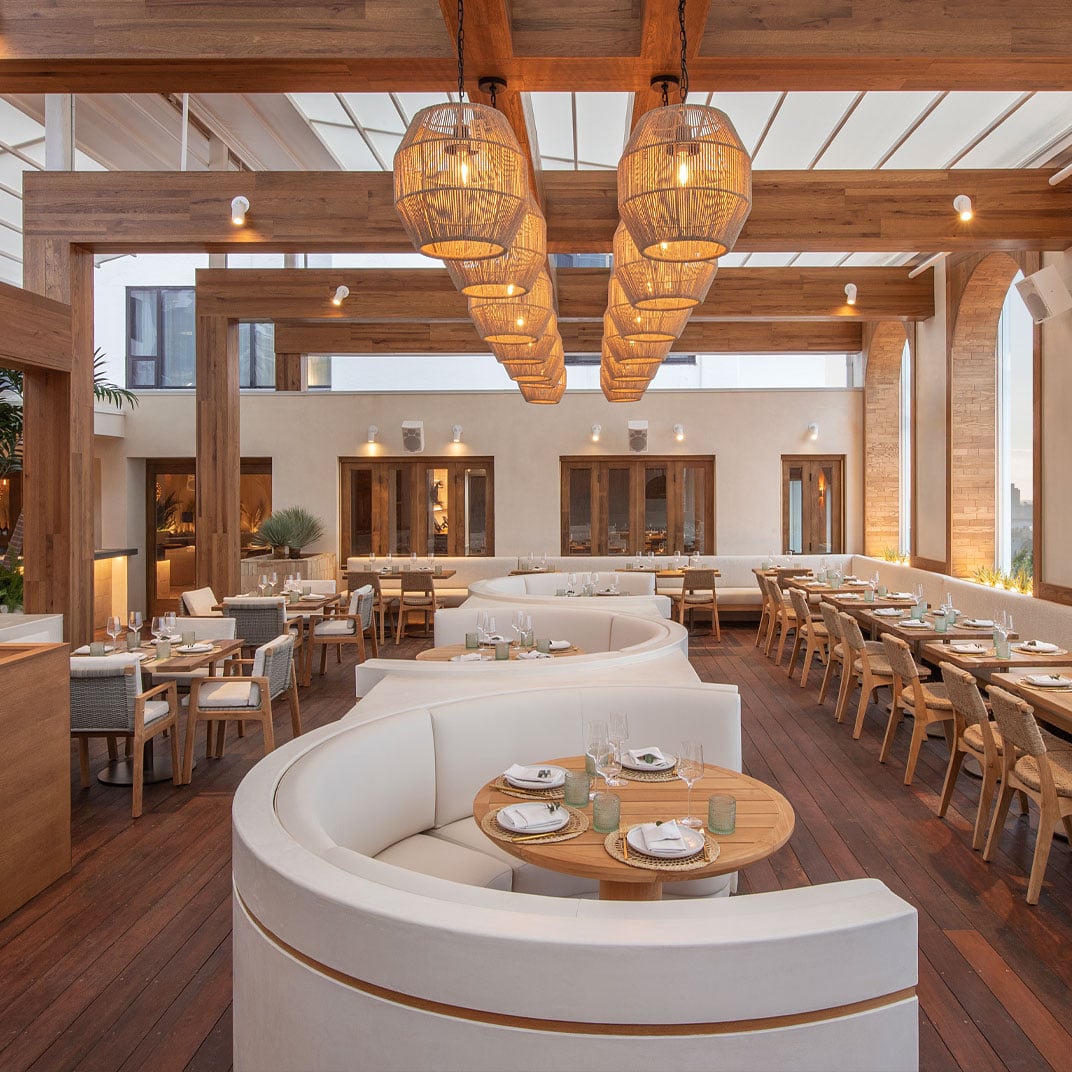 Wicker pendant lights cast warm shadows over curved white banquettes and honey-toned wood, creating an effortlessly chic dining atmosphere beneath the open pergola ceiling.