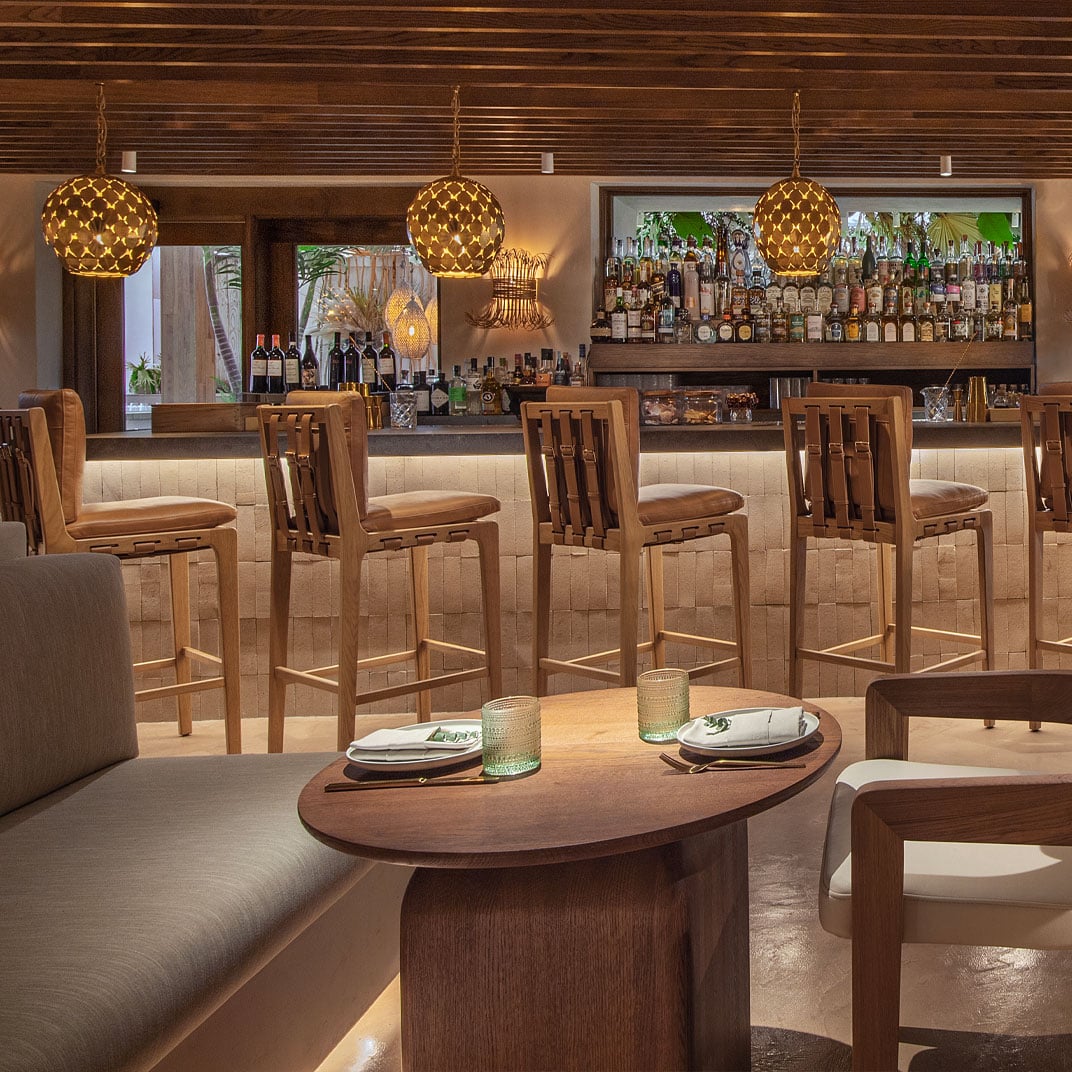 Woven pendant lights cast golden warmth over the sleek bar where craft cocktails flow beneath a striking wood-slat ceiling and verdant glimpses of West Hollywood.
