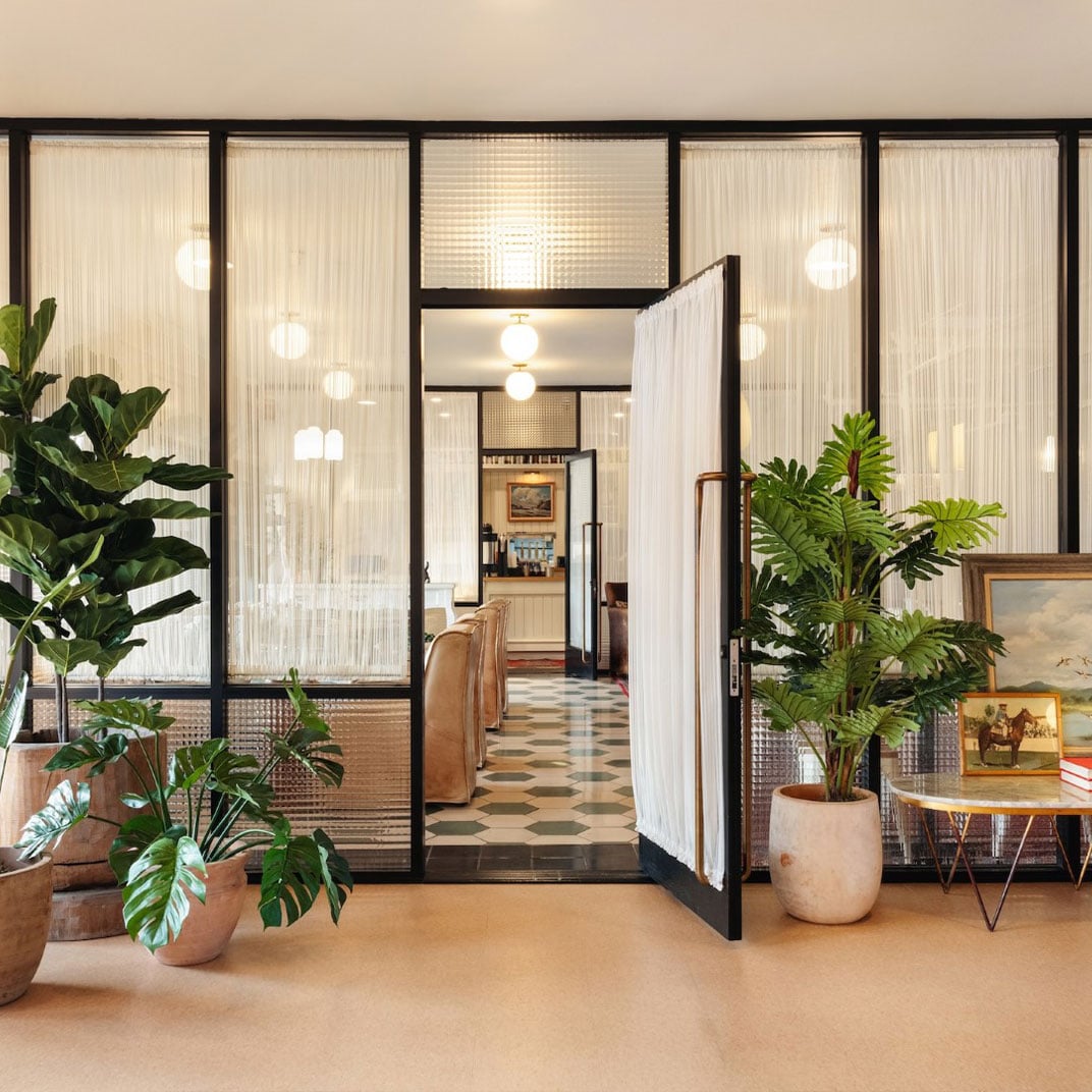 Fluted glass partitions and sprawling greenery create an inviting threshold between public and private spaces, where warm pendant lights cast a golden glow over mid-century furnishings.