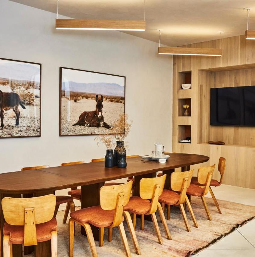 Warm wood tones and desert photography create an intimate dining atmosphere, where handcrafted chairs gather around a sculptural walnut table beneath glowing linear pendants.