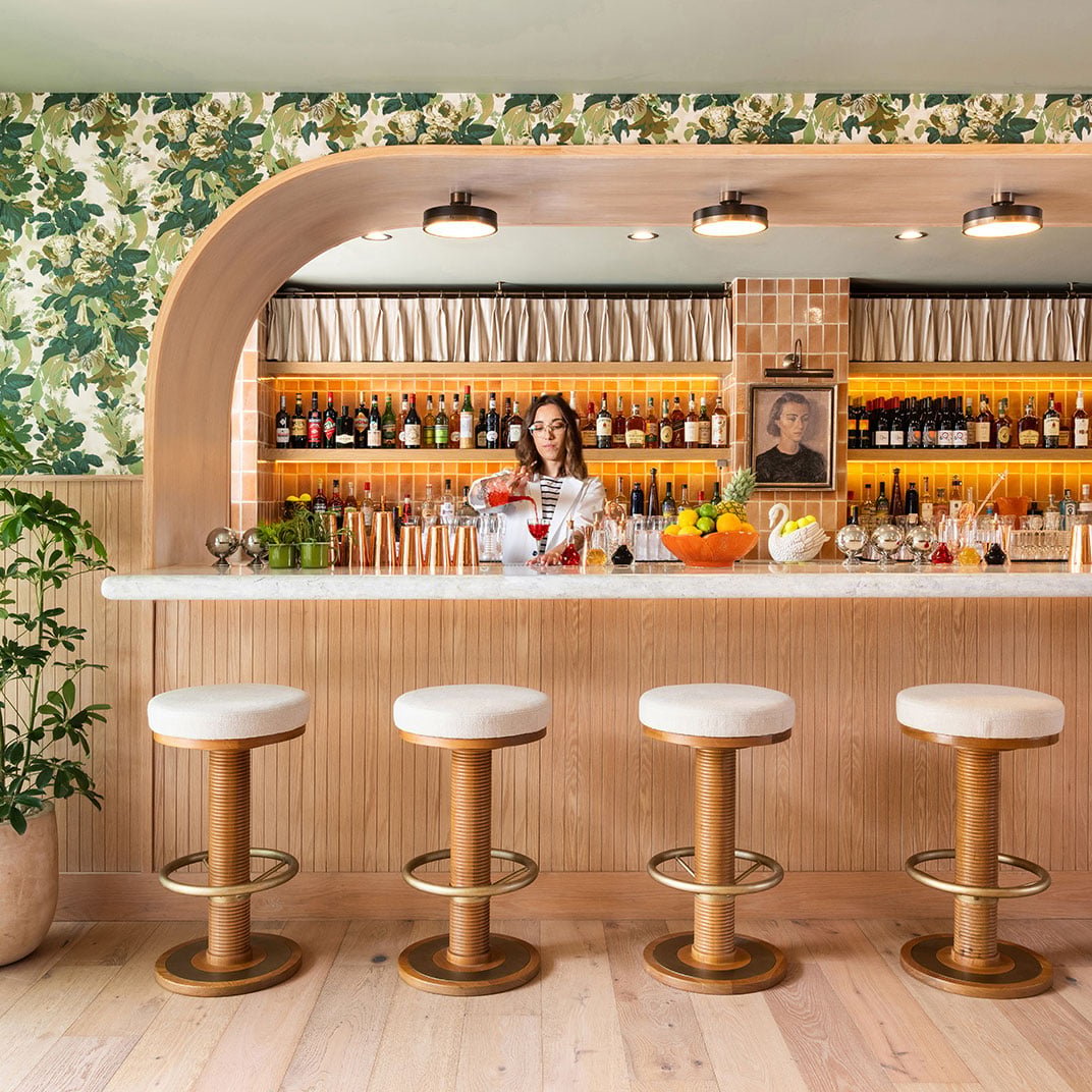Botanical wallpaper and brass-legged stools create an intimate cocktail sanctuary where skilled bartenders craft drinks beneath warm pendant lighting and vintage portraiture.