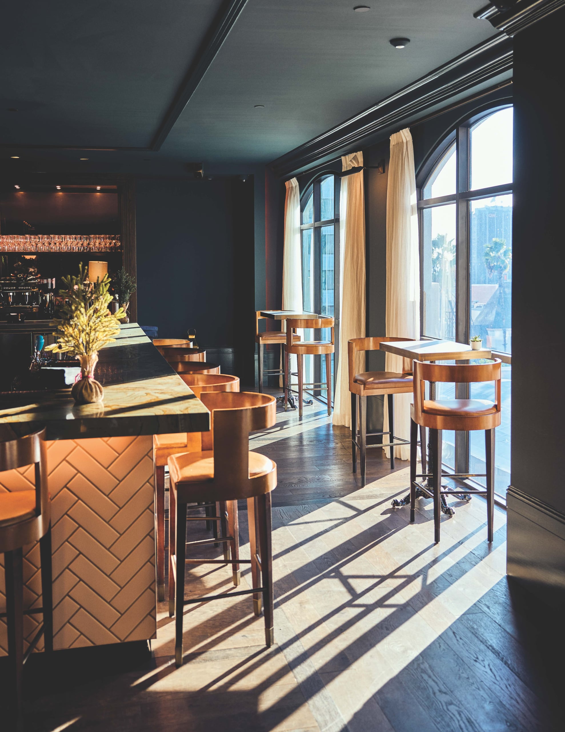 Warm leather bar stools catch afternoon light streaming through tall windows, creating intimate shadows across polished wood floors in this sophisticated drinking den.