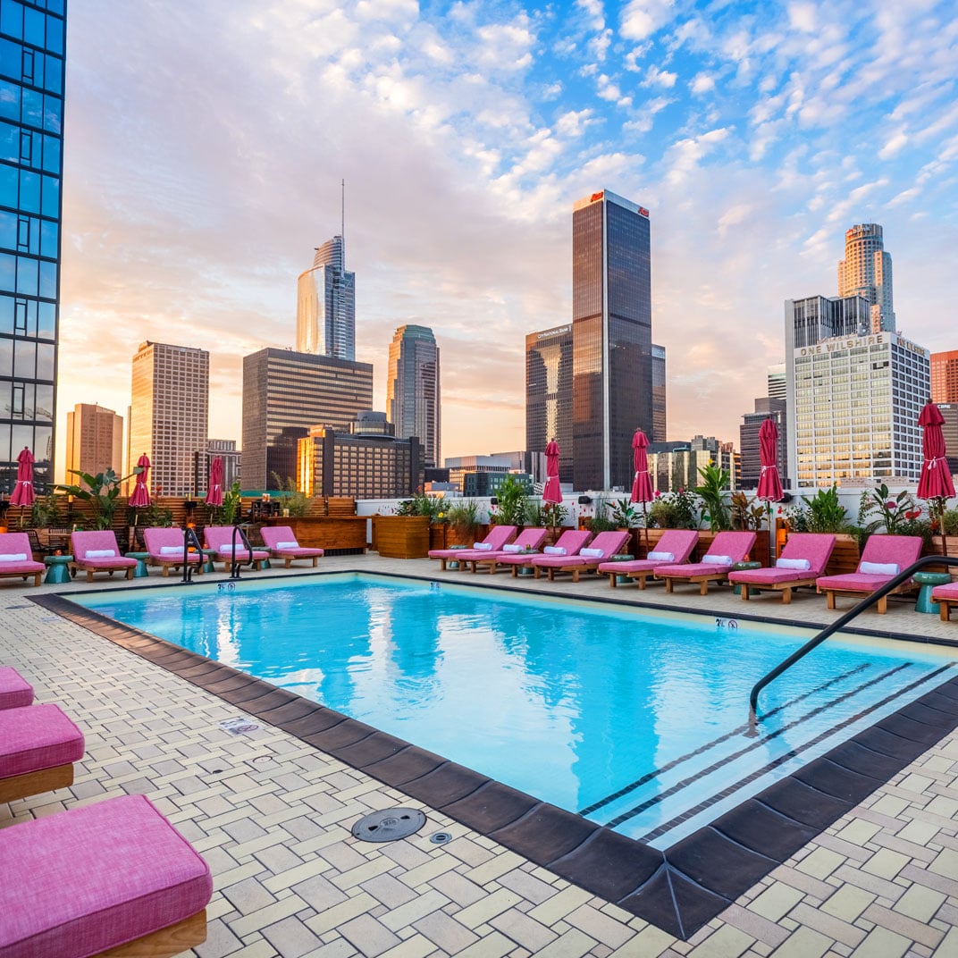 Pink-cushioned loungers frame the rooftop pool as downtown LA's gleaming towers catch golden hour light, creating an urban oasis above the bustling streets below.
