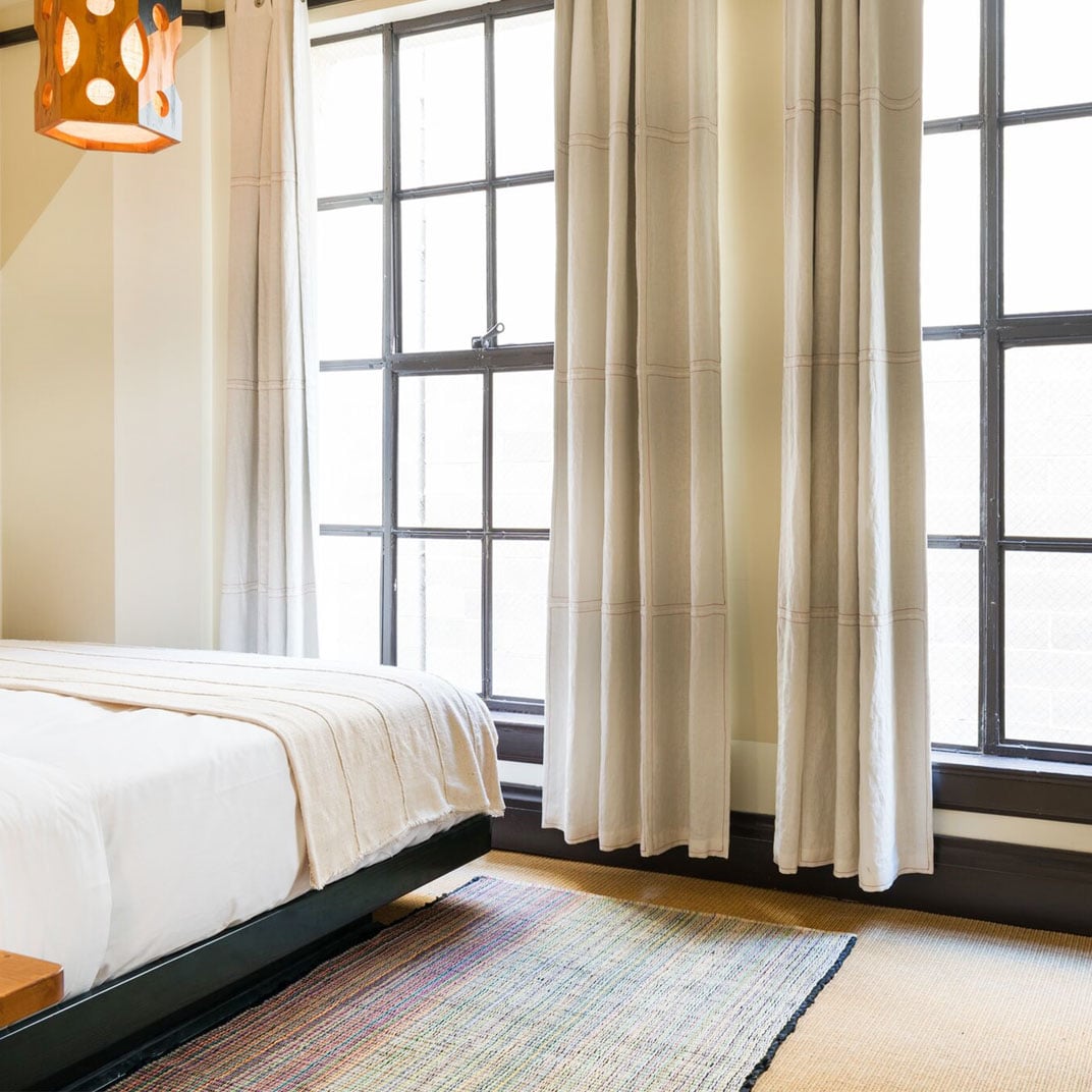 Natural light floods through industrial-style windows, illuminating crisp linens and a handwoven rug that adds warmth to minimalist accommodations with mid-century touches.
