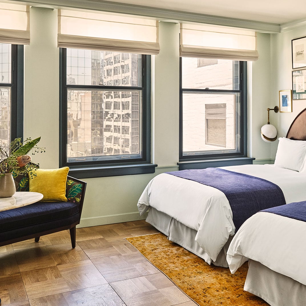 Twin beds dressed in crisp white linens and navy runners create a serene sanctuary, while floor-to-ceiling windows frame the bustling cityscape beyond.