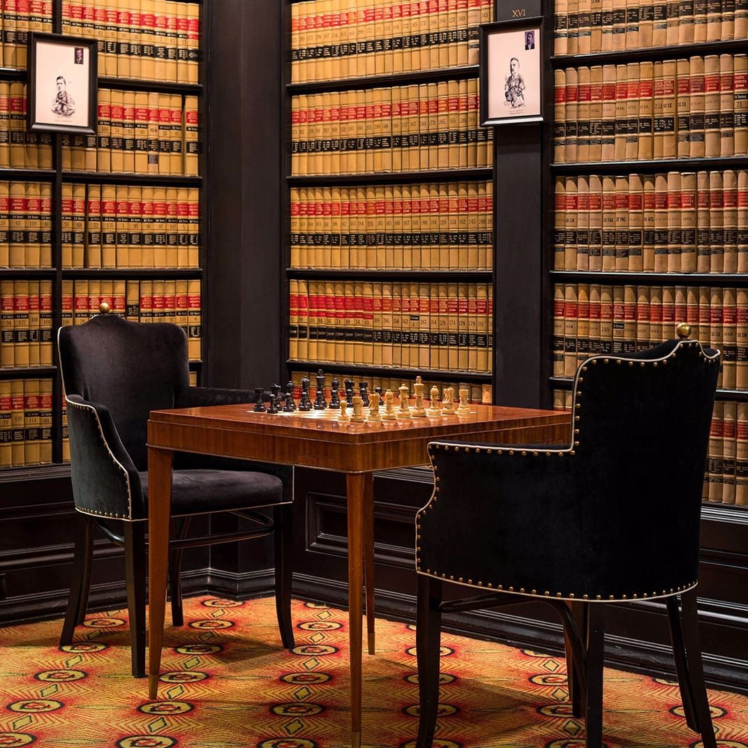Floor-to-ceiling law books create an intimate library corner where guests settle into leather chairs for chess, surrounded by golden volumes and vintage portraits.