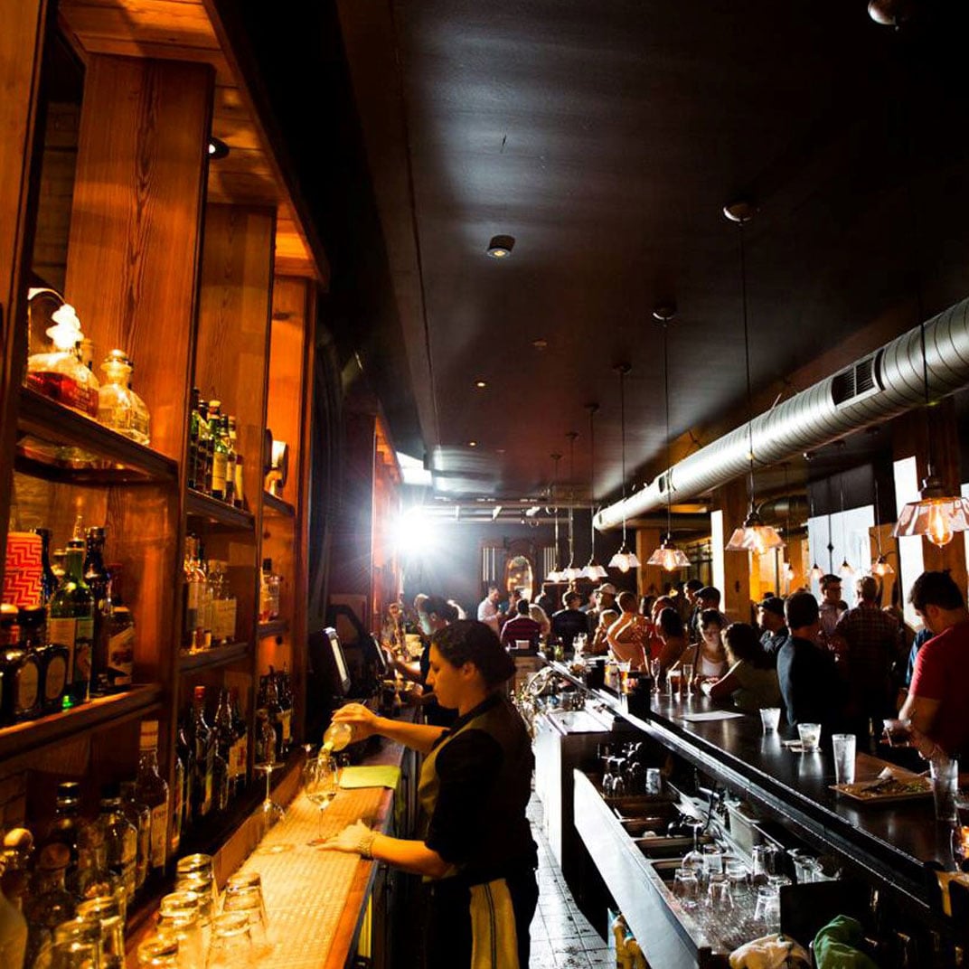 Exposed ductwork and warm wood shelving create industrial elegance as bartenders craft cocktails for a lively crowd beneath moody pendant lighting.