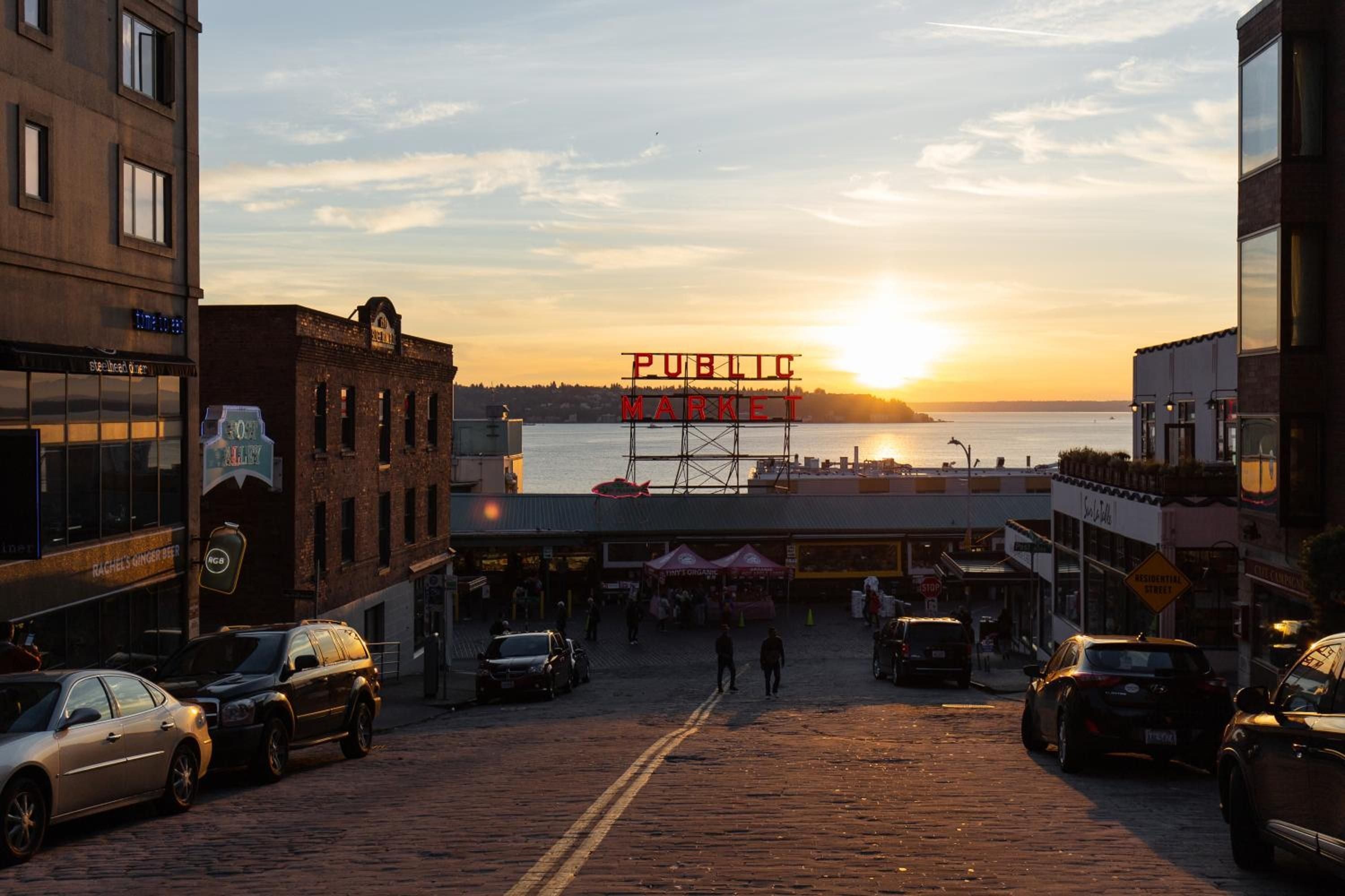 Golden hour light bathes Pike Place Market's iconic neon sign as the sun sets over Elliott Bay, framing Seattle's most beloved waterfront destination in warm amber hues.