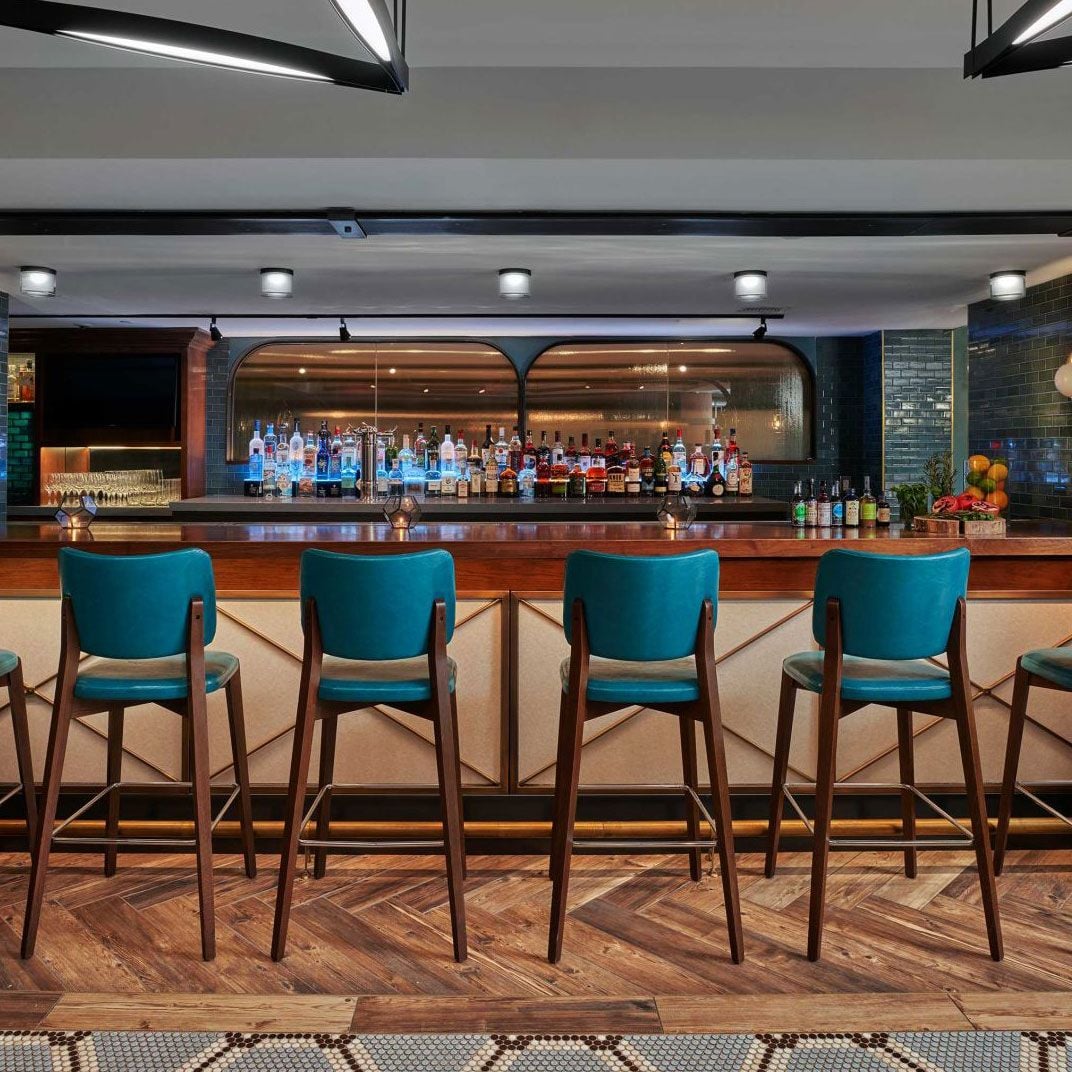 Teal leather stools line the polished wood bar where gleaming bottles catch the light beneath geometric archways, creating an intimate mid-century modern drinking den.