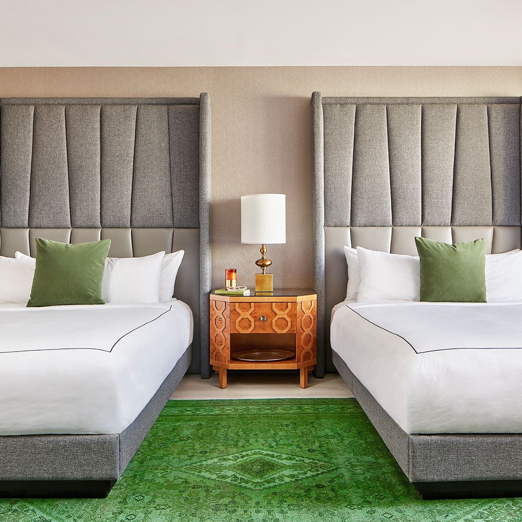 Twin beds adorned with sage velvet pillows flank a geometric wood nightstand, while an emerald rug anchors this sophisticated guest room's earthy palette.