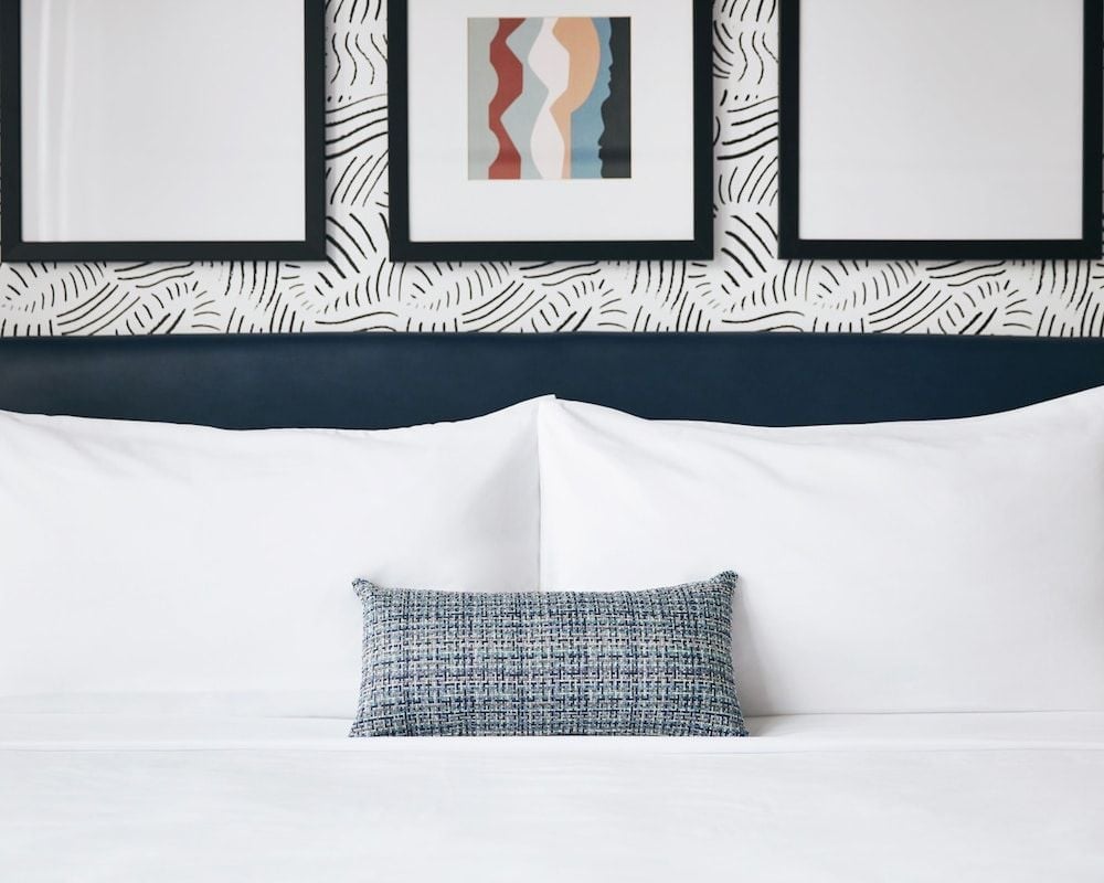 Crisp white linens meet playful palm-print wallpaper in thoughtfully appointed guest rooms, where textured throw pillows and curated artwork create an inviting urban retreat.