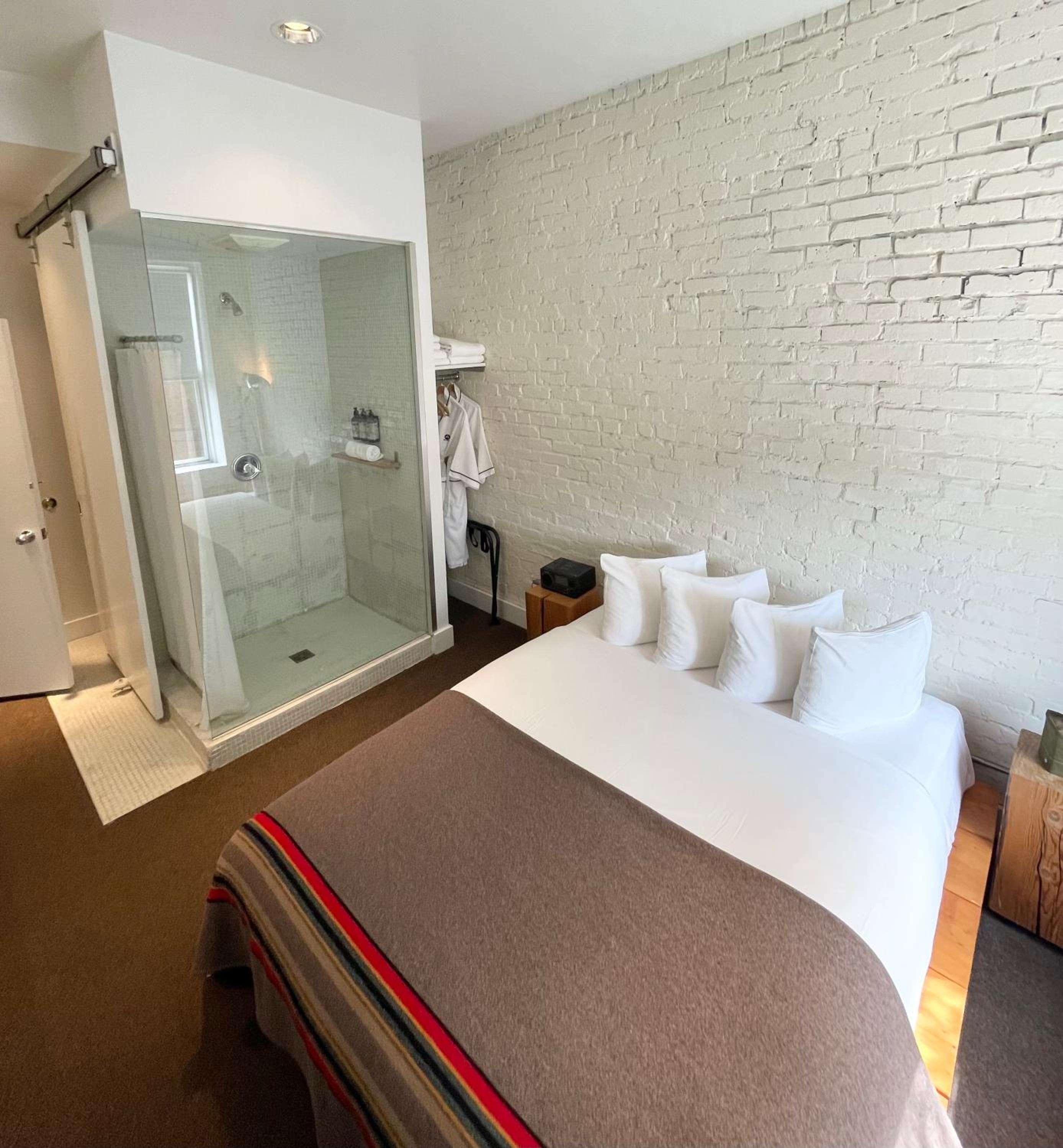 Exposed white brick walls and crisp linens create an industrial-chic sanctuary, while glass-enclosed showers blur the boundaries between sleeping and bathing spaces.