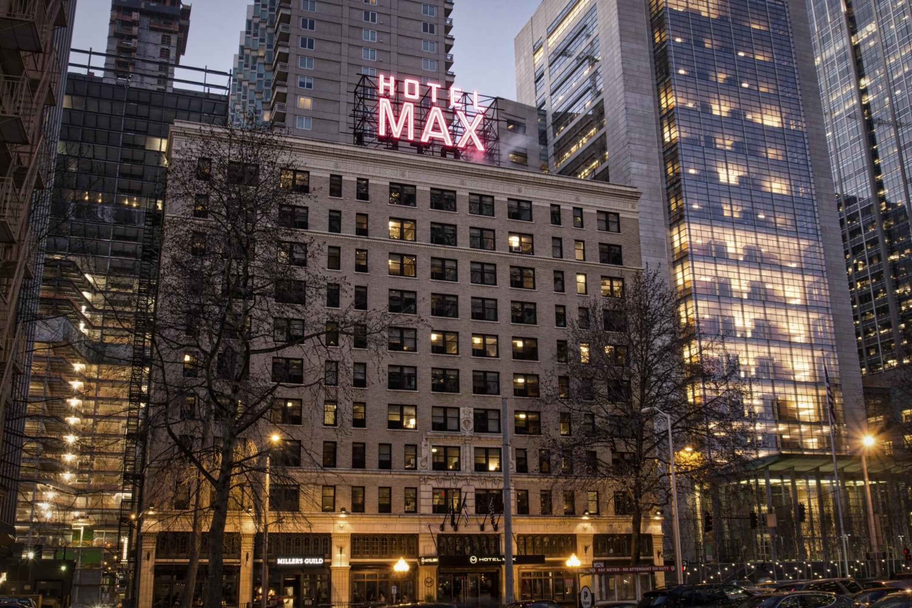 Vintage brick facade meets modern Seattle skyline as Hotel Max's glowing rooftop sign cuts through twilight, while street-level warmth spills from illuminated storefronts below.