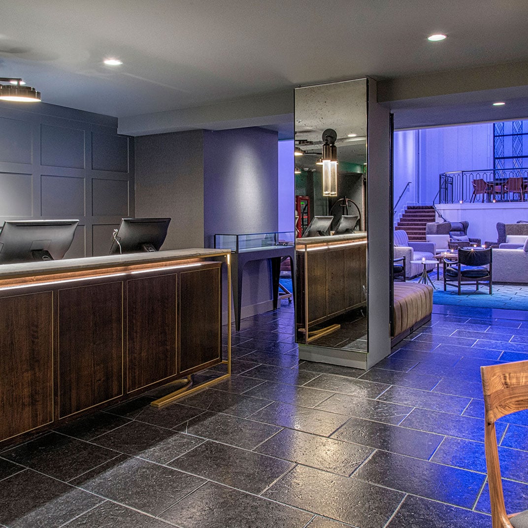 Warm walnut cabinetry and cool blue lighting create sophisticated contrasts in this modern lobby space, where sleek surfaces meet inviting lounge areas beyond.