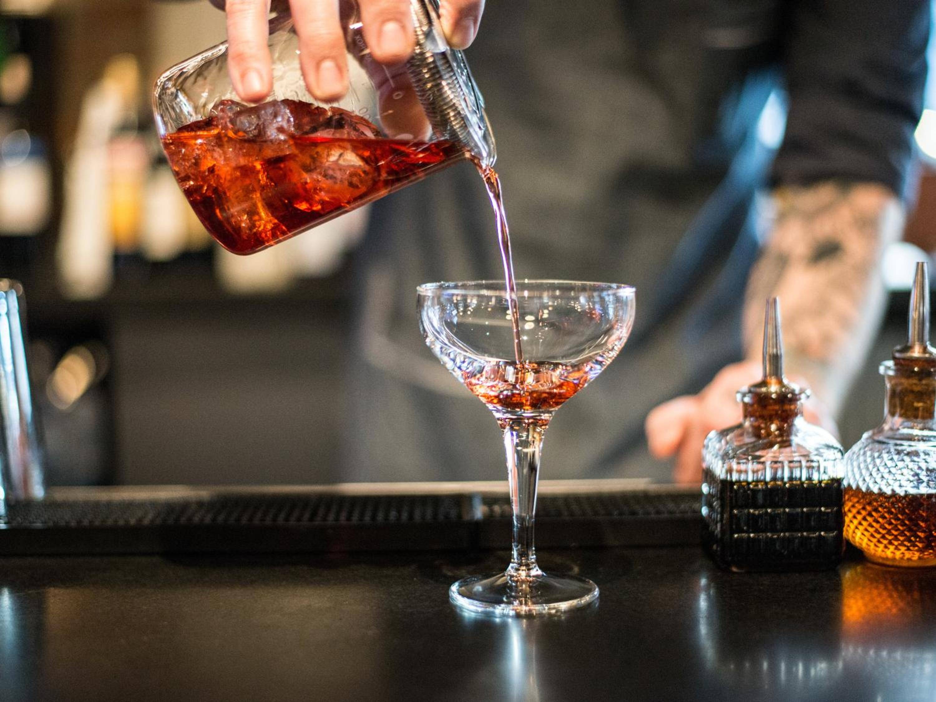 Amber spirits cascade from crystal mixing glass to coupe at Hotel Theodore's sophisticated bar, where classic cocktail artistry unfolds with theatrical precision and golden light.