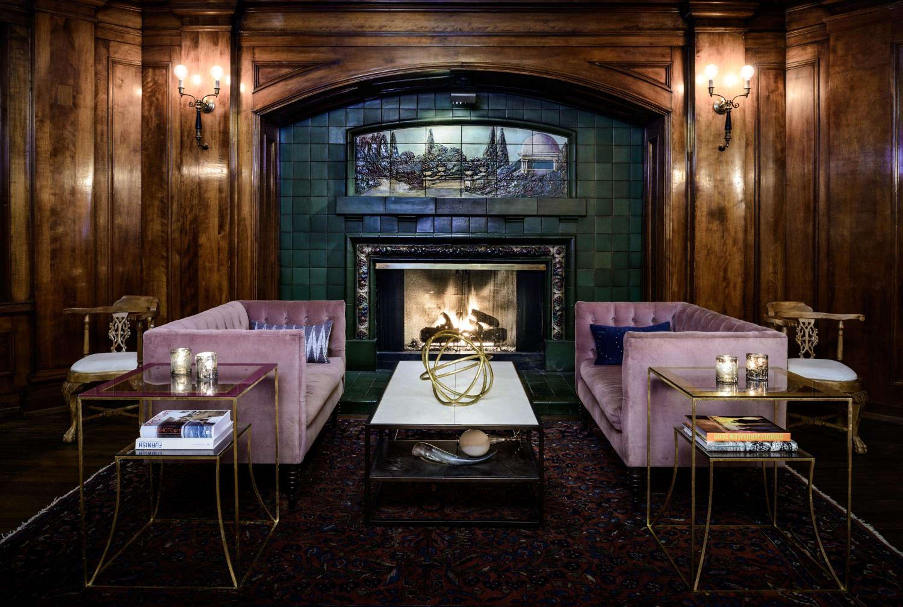 Velvet sofas in dusty rose face a crackling fireplace beneath ornate green tiles, where warm sconces cast intimate shadows across rich walnut paneling.