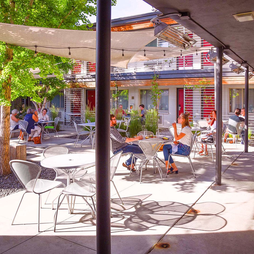 Sunlight filters through the covered terrace where guests gather at sleek metal tables, creating an inviting communal atmosphere beneath modern red-accented architecture.
