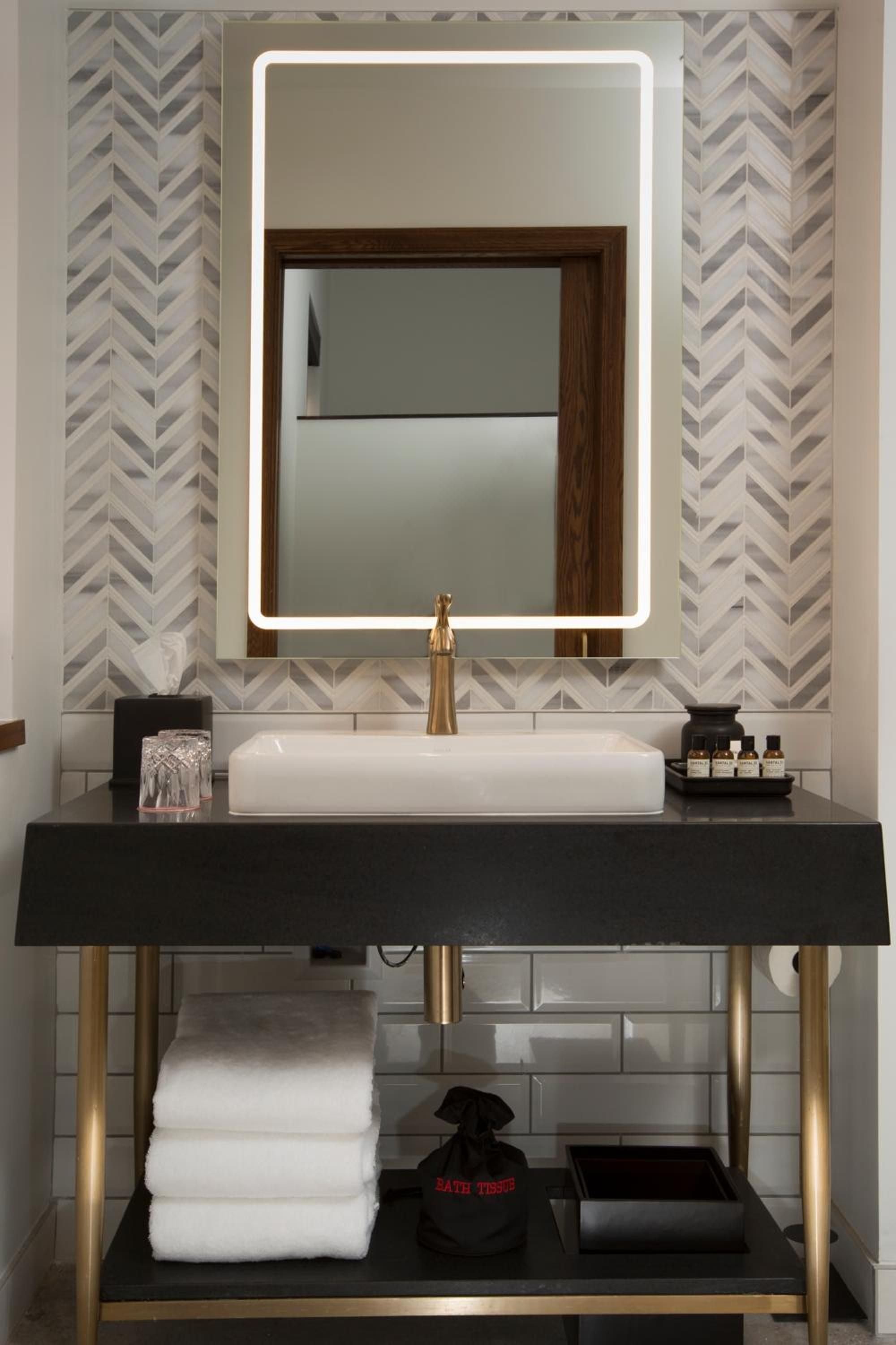 Brass fixtures gleam against chevron marble while LED lighting frames the vanity mirror, creating a sophisticated interplay of warm metals and cool stone textures.